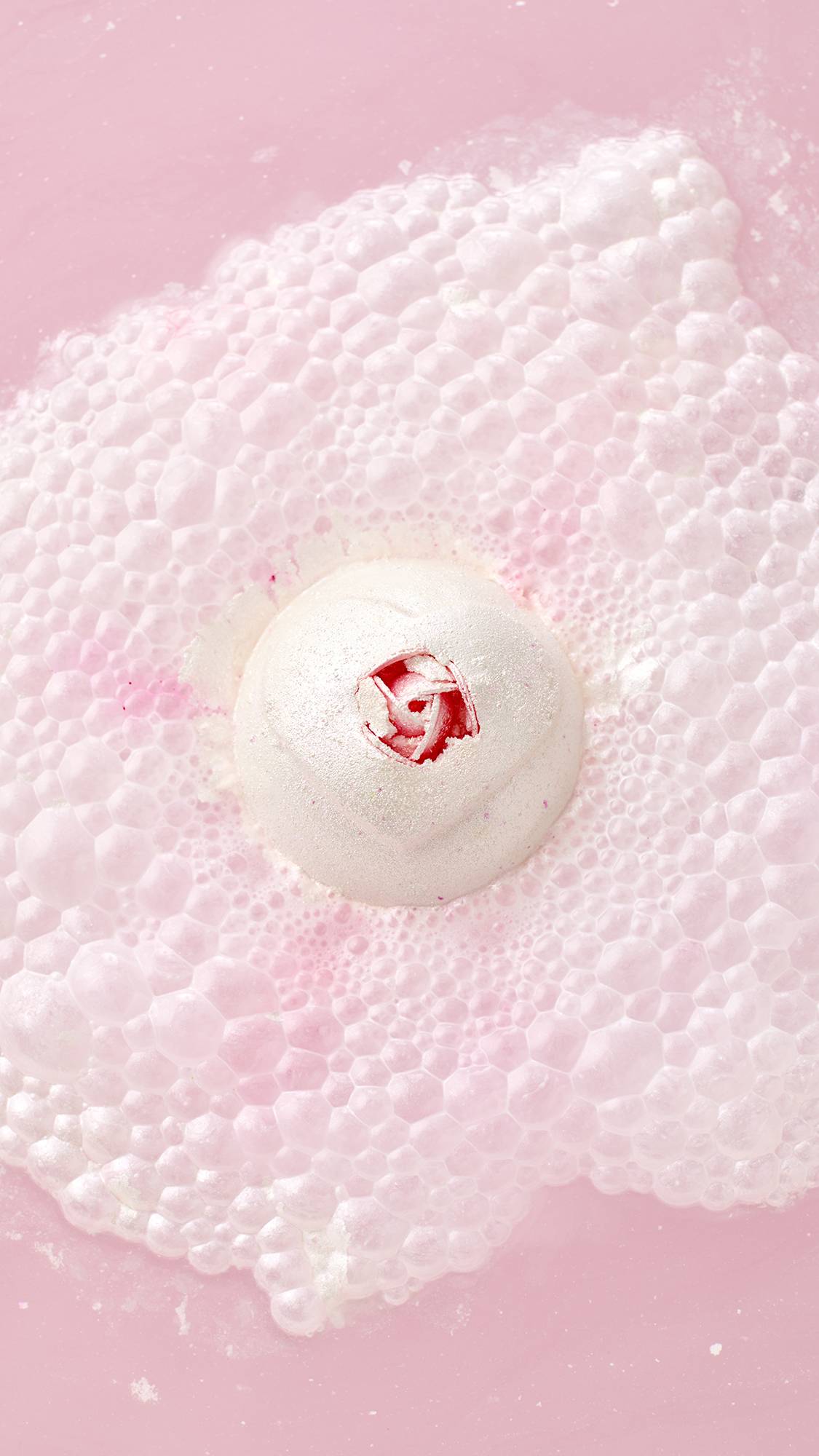 A top-down view of the white Wedding bath bomb with a pink rose center, floating in pink water. It generates a massive, textural cloud of white and pink bubbles and foam.