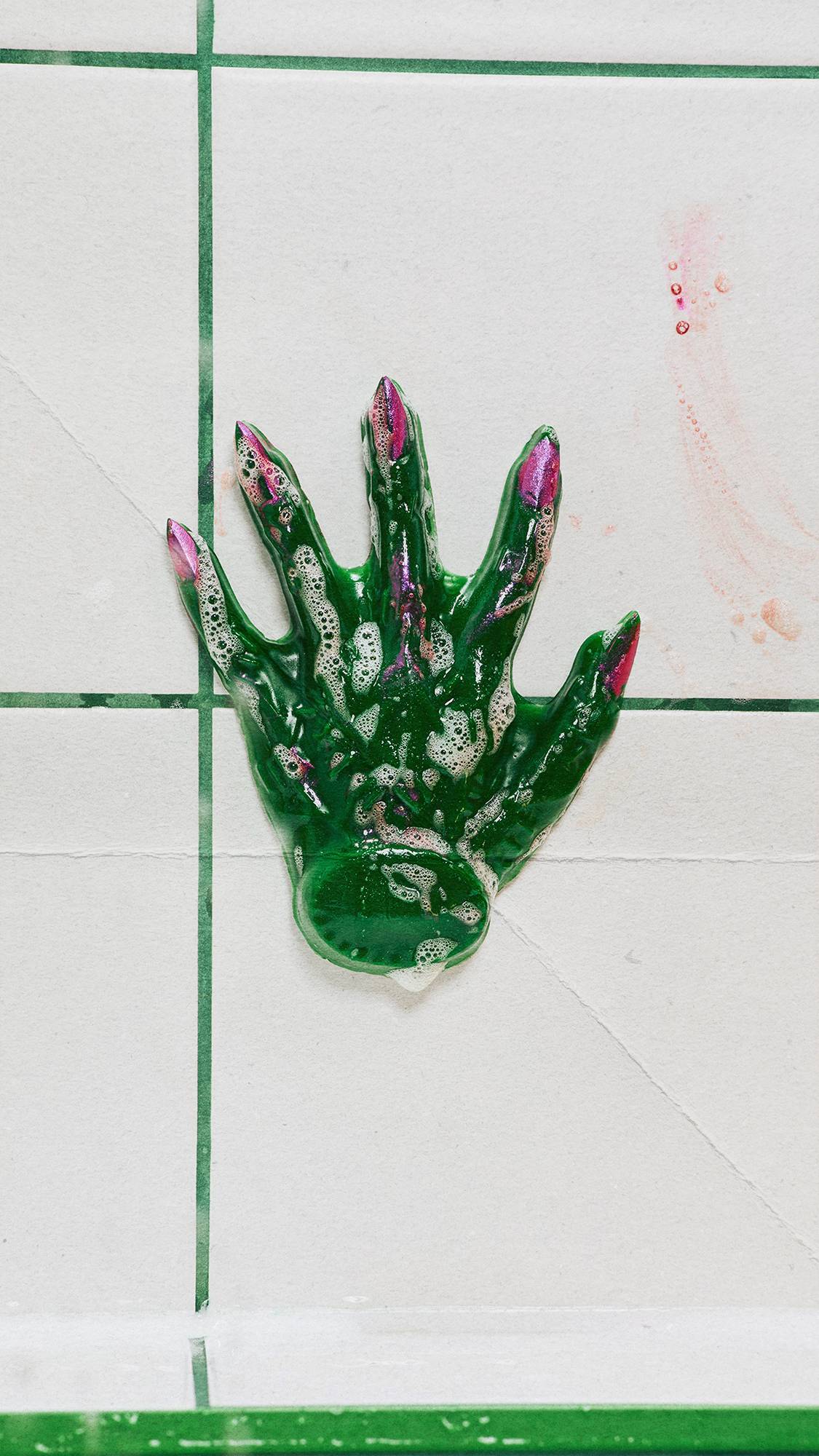 A dark green shower gummy shaped like a witch's hand with metallic pink fingernails stuck onto a wet shower wall. Small patches of white soap foam are visible on its surface. White square wall tiles with green grout form the background.