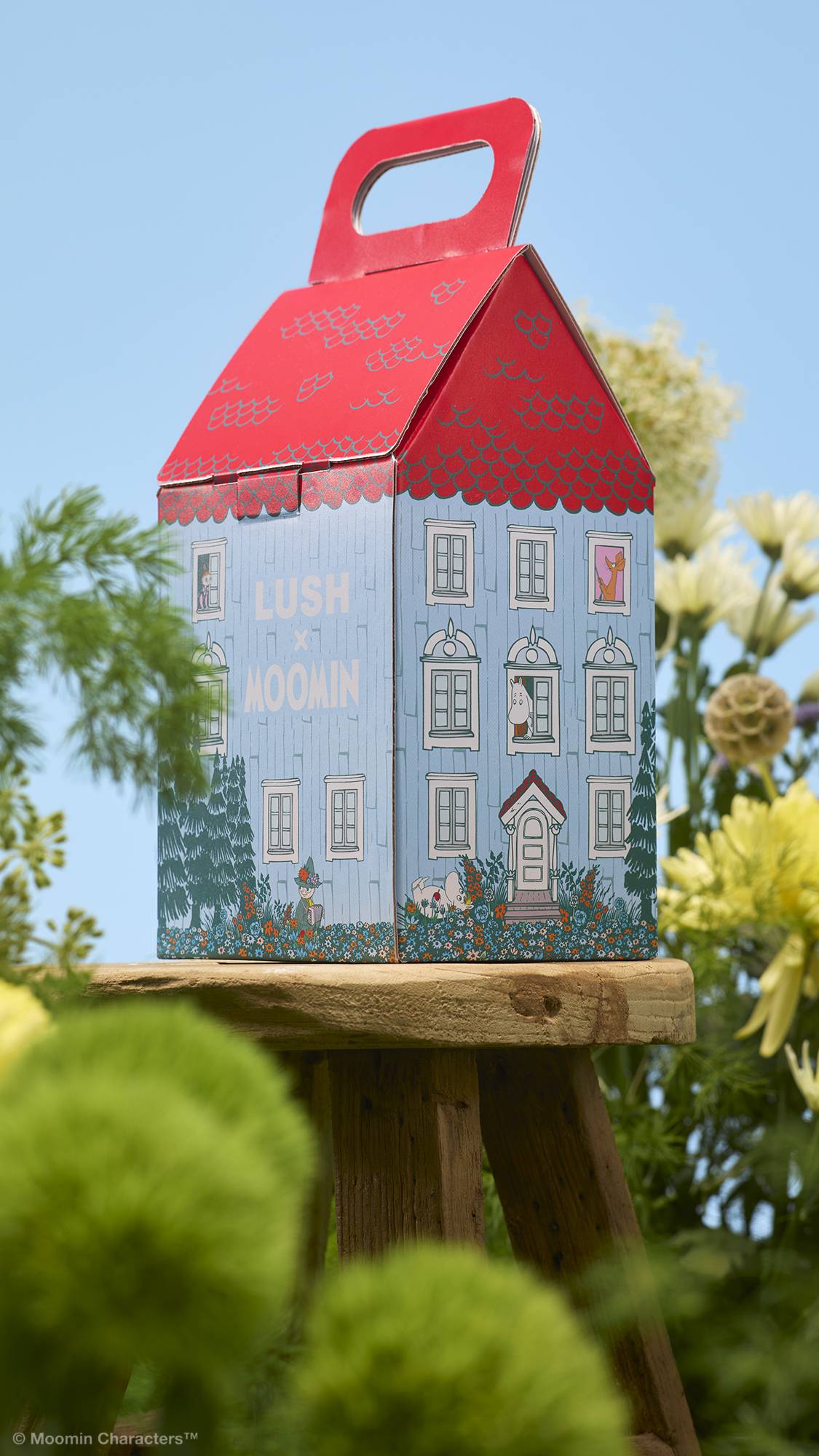 A house-shaped "LUSH x MOOMIN" gift box sits on a small wooden stool. The box is illustrated to look like a blue house with a red roof. Green foliage and yellow flowers frame the stool against a clear blue sky.