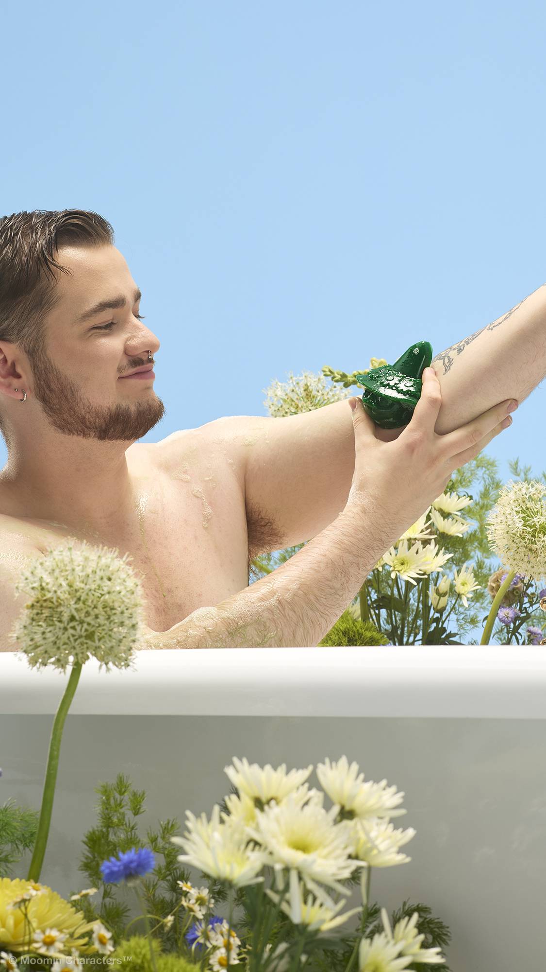 A close-up shot of a person with a beard sitting in a white bathtub, washing their arm with a green Snufkin-shaped shower gummy. Yellow and white wildflowers surround the tub against a clear blue sky background.
