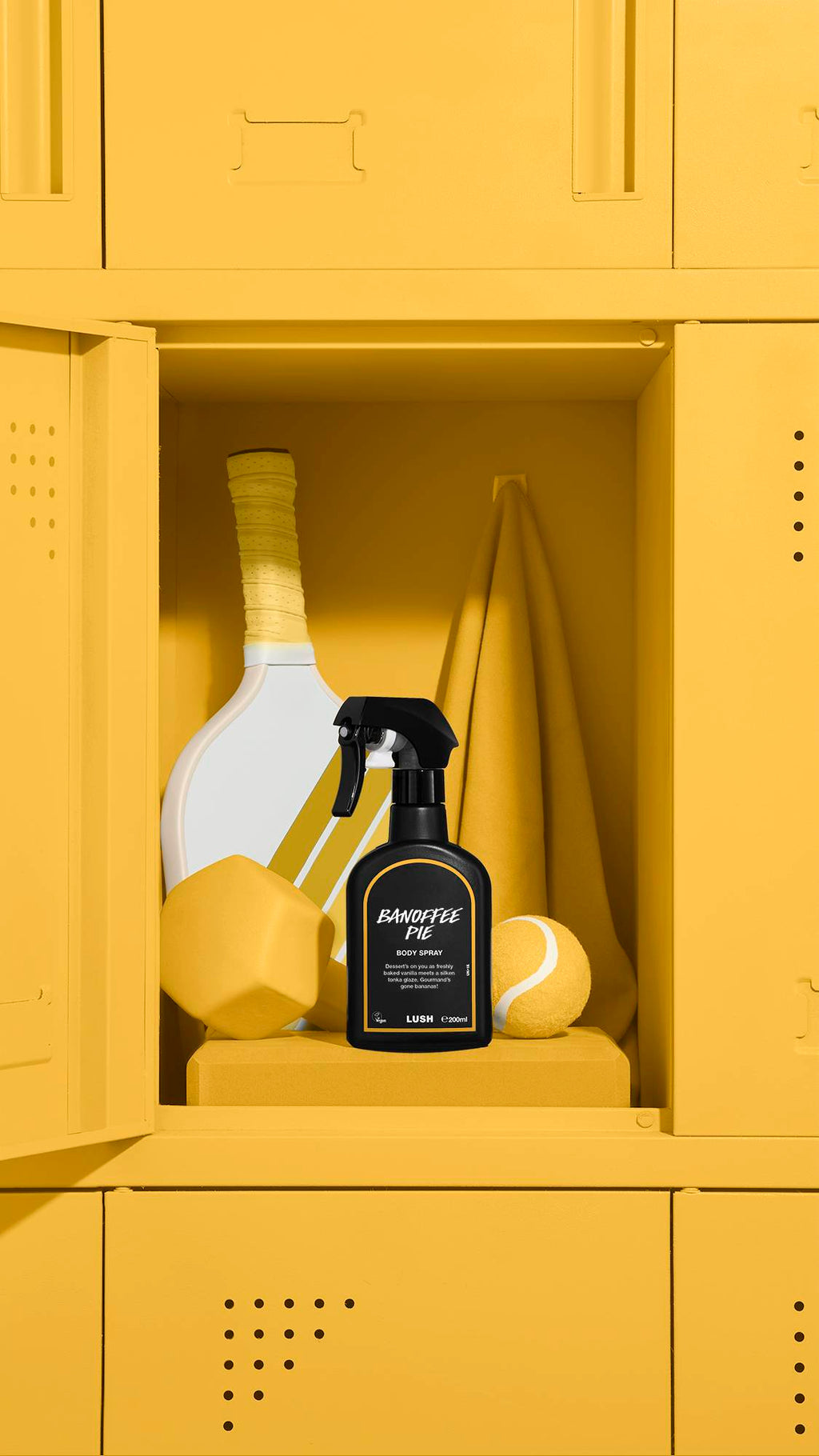 A black bottle of Lush 'Banoffee Pie' body spray sits inside an open yellow locker. Behind it are a matching yellow towel, a tennis racket, a ball, and a dumbbell. The entire locker unit and all sports equipment are a uniform bright yellow.