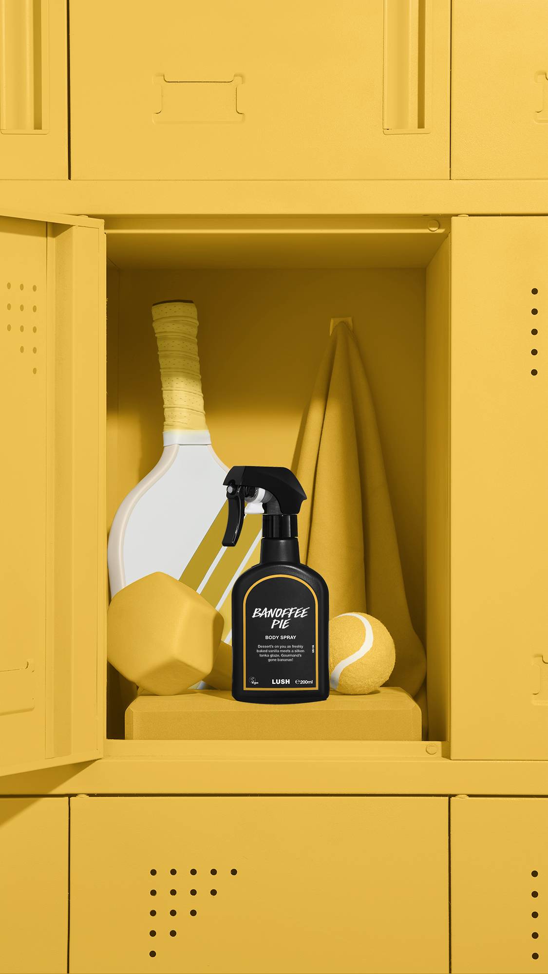 A black bottle of Lush 'Banoffee Pie' body spray sits inside an open yellow locker. Behind it are a matching yellow towel, a tennis racket, a ball, and a dumbbell. The entire locker unit and all sports equipment are a uniform bright yellow.