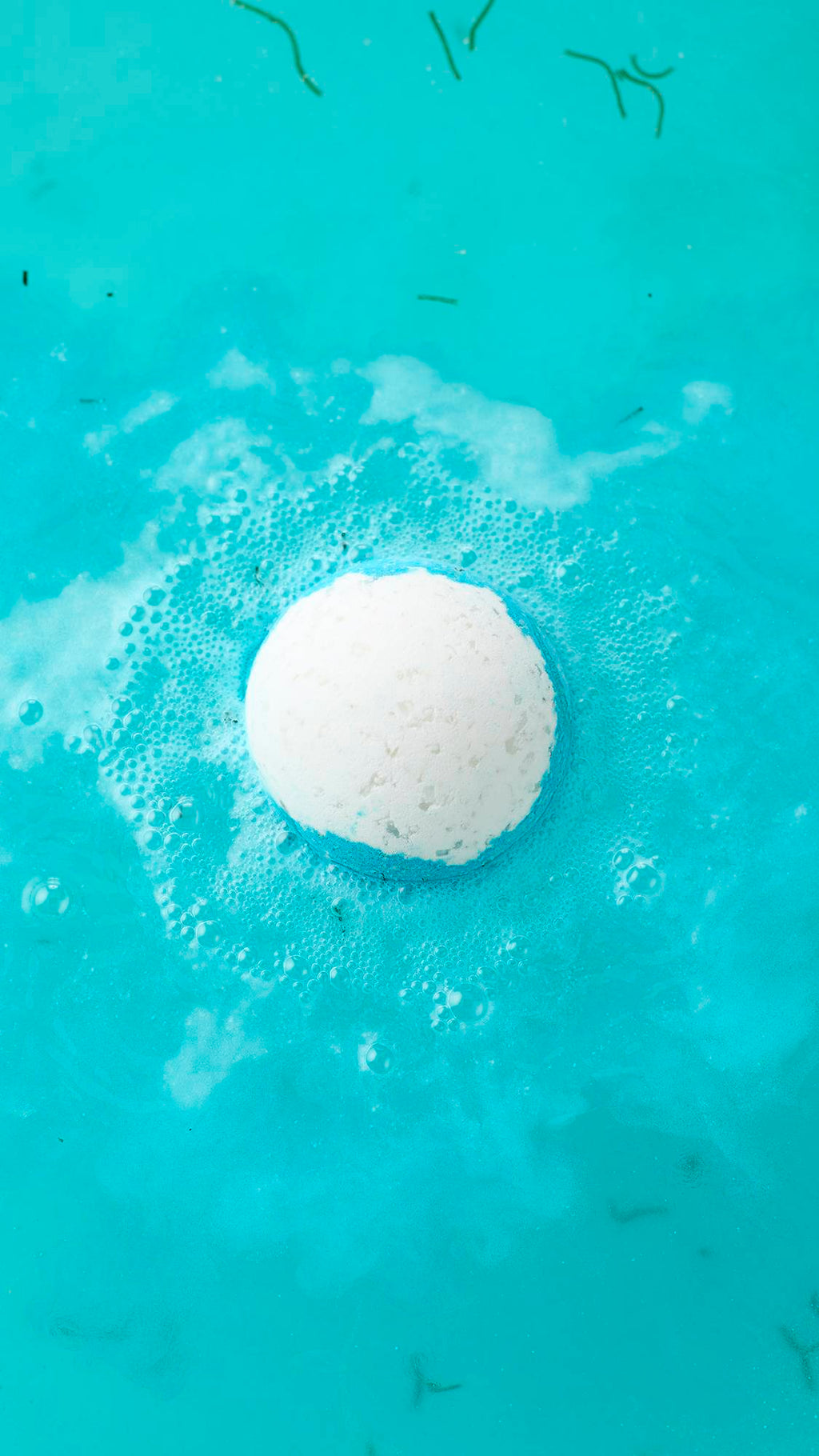 A top-down view of the Big Blue bath bomb fizzing in turquoise water. The white sphere is surrounded by a halo of blue and white foam, with small green seaweed strands floating in the vibrant bathwater.