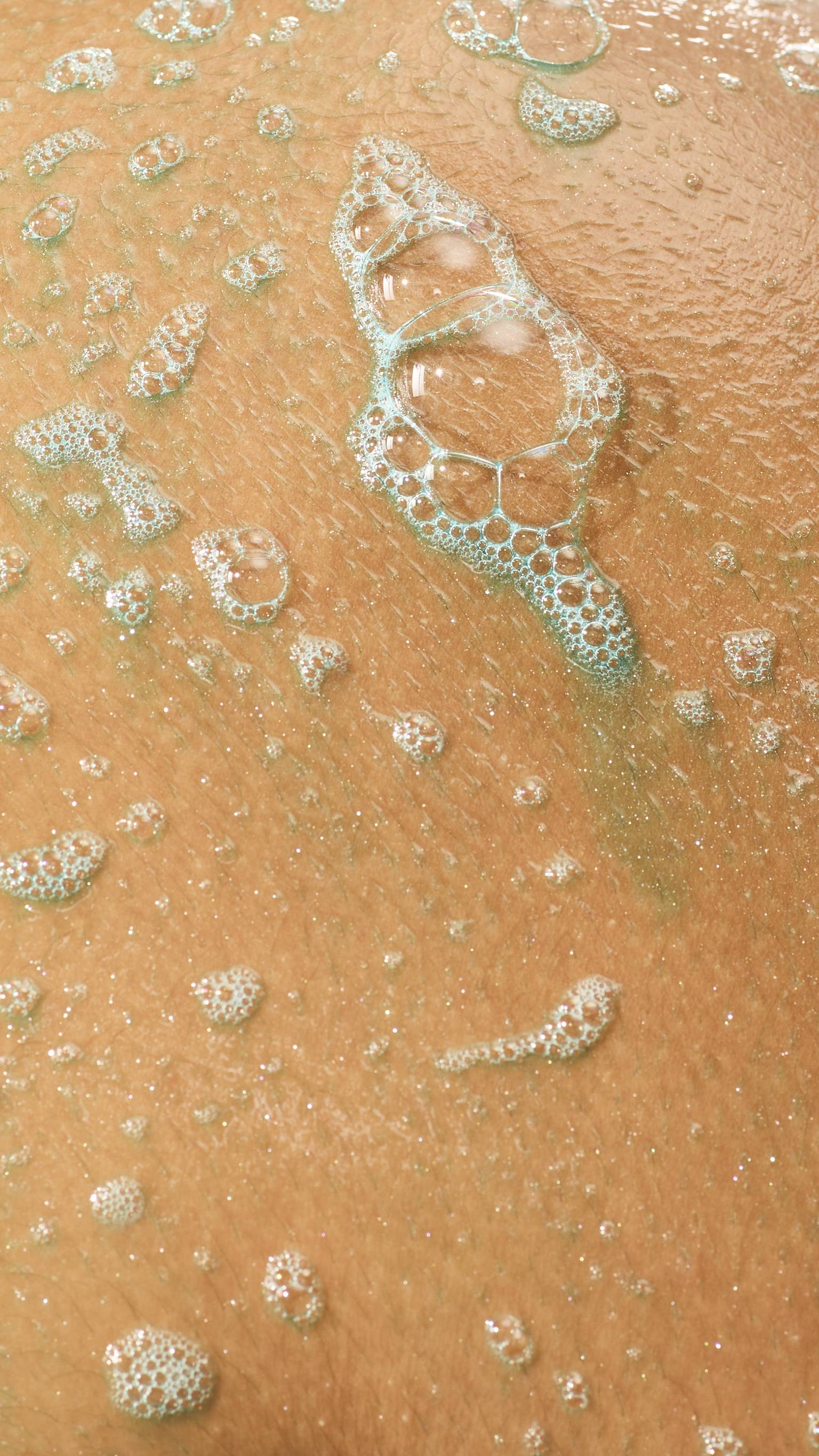 A close-up of wet, glistening skin covered in light teal soapy lather and clusters of translucent bubbles.