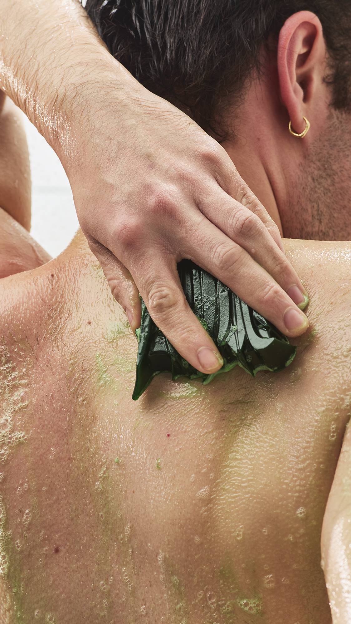 A close-up view from behind of a person rubbing a textured, dark green shower gummy soap across their wet back, creating a light green lather with soapy bubbles.