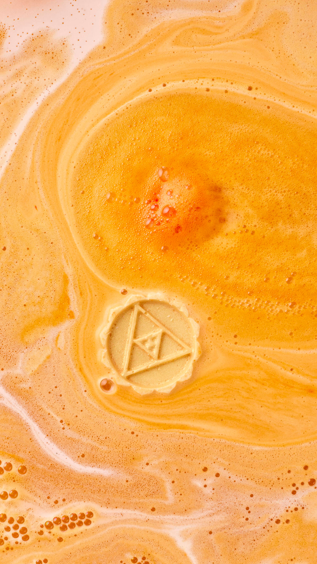 An aerial view of the bathwater after using the Karma macaron-shaped bath bomb. The orange product has dissolved, leaving spiralling swirls of thick, velvety, golden foam. The solid oil central layer floats, slowly dissolving.
