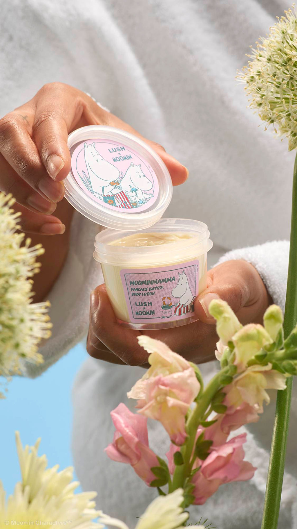 A close-up shot of hands holding open a clear pot of cream-coloured "Moominmamma Pancake Batter" body lotion. The pink labels feature Moomin illustrations, framed in the foreground by soft pink and white wildflowers.