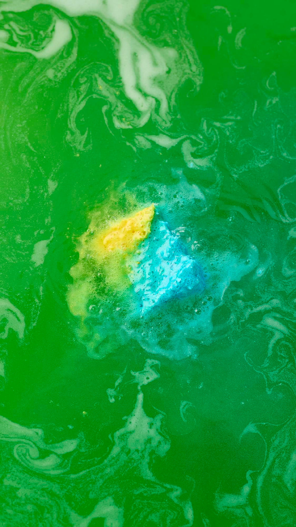 An aerial view of the blue and yellow cube-shaped "Salt Water Soother" bath bomb dissolving in vibrant green water. It is surrounded by frothy bubbles, with streaks of blue, yellow, and white foam spiralling outwards into the green liquid.