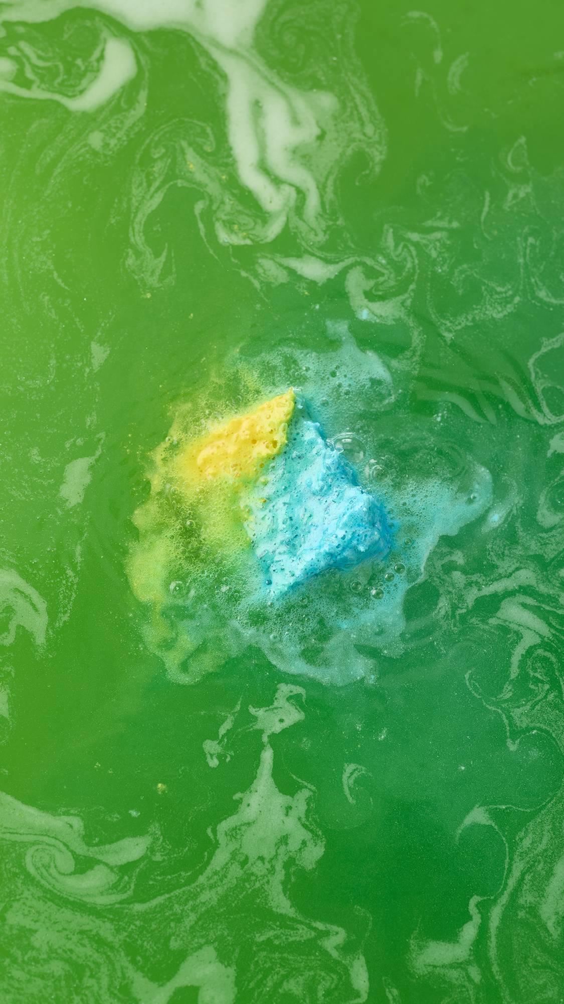 An aerial view of the blue and yellow cube-shaped "Salt Water Soother" bath bomb dissolving in vibrant green water. It is surrounded by frothy bubbles, with streaks of blue, yellow, and white foam spiralling outwards into the green liquid.
