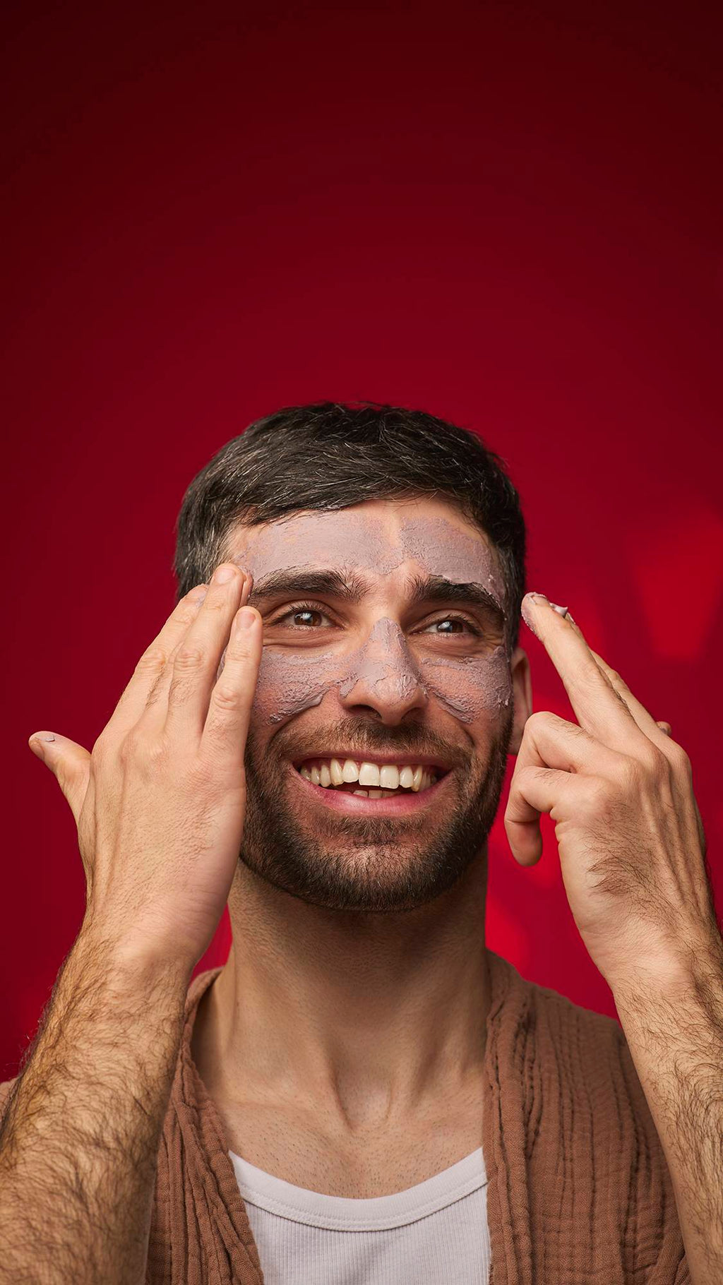 A smiling person applies the mauve-toned Sipak face mask to their forehead and nose. They wear a brown textured robe against a solid deep red background.