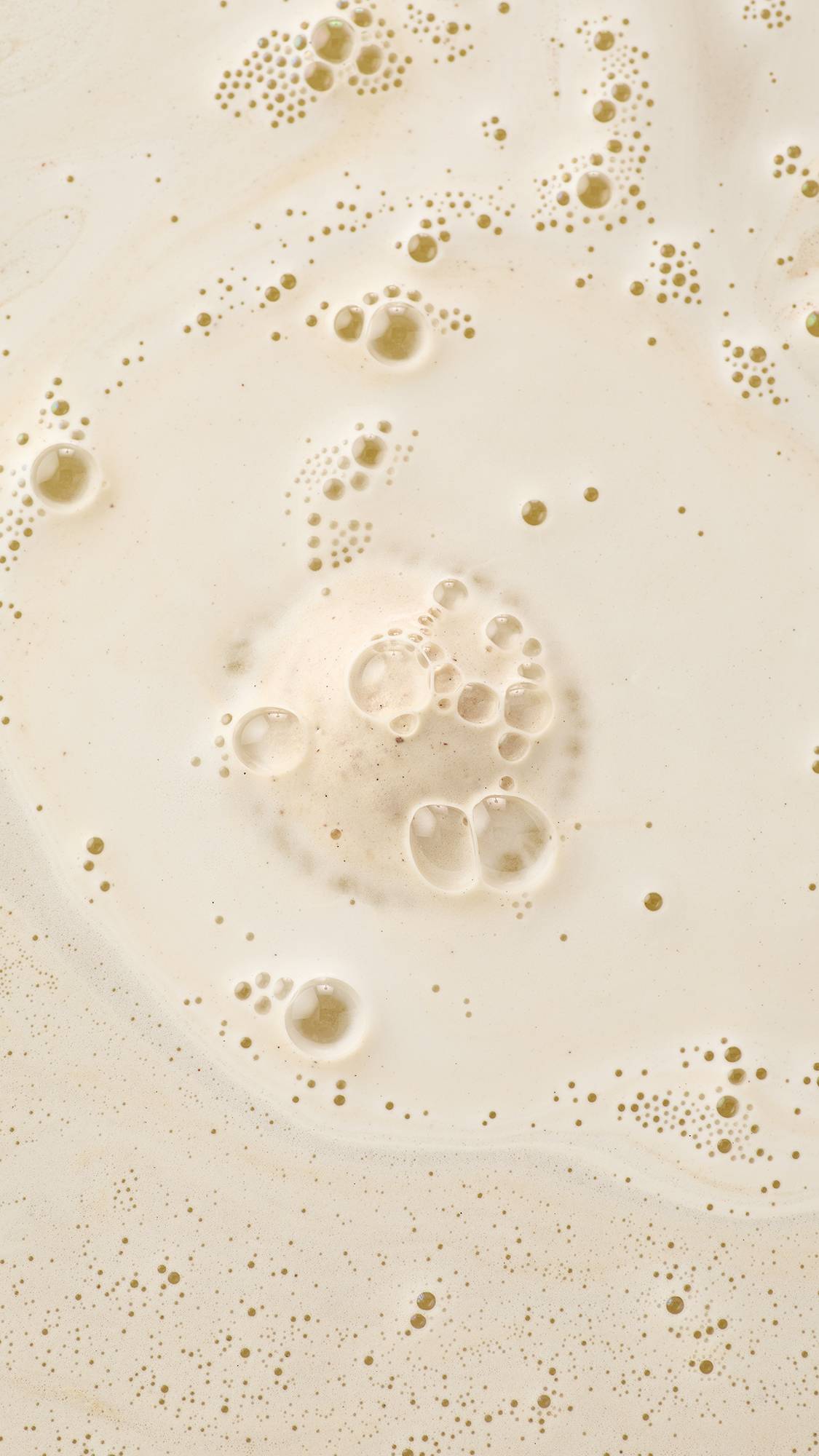 An aerial, abstract view of creamy bathwater. Fine, frothy bubbles and larger translucent suds cluster and swirl across the surface, creating a marbled and textured effect of velvety foam.