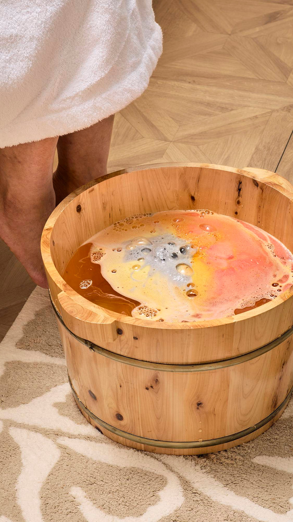 The image shows the "Volcano" foot soak as it dissolves in a wooden foot tub, turning the water a deep golden-orange. Swirls of grey, yellow, and pink foam spread across the surface, creating a colourful, bubbling display as it fizzes away.