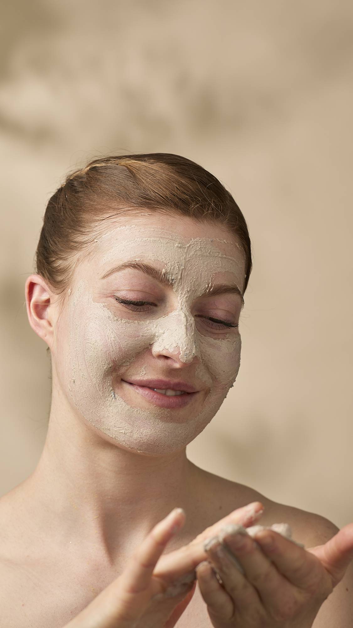 A model with their hair tied back looks down and smiles as they apply a thick, beige "Tiny Musicians Of The Forest" facial cleanser. The creamy, textured mask covers most of their face as they rub more of the product between their hands.