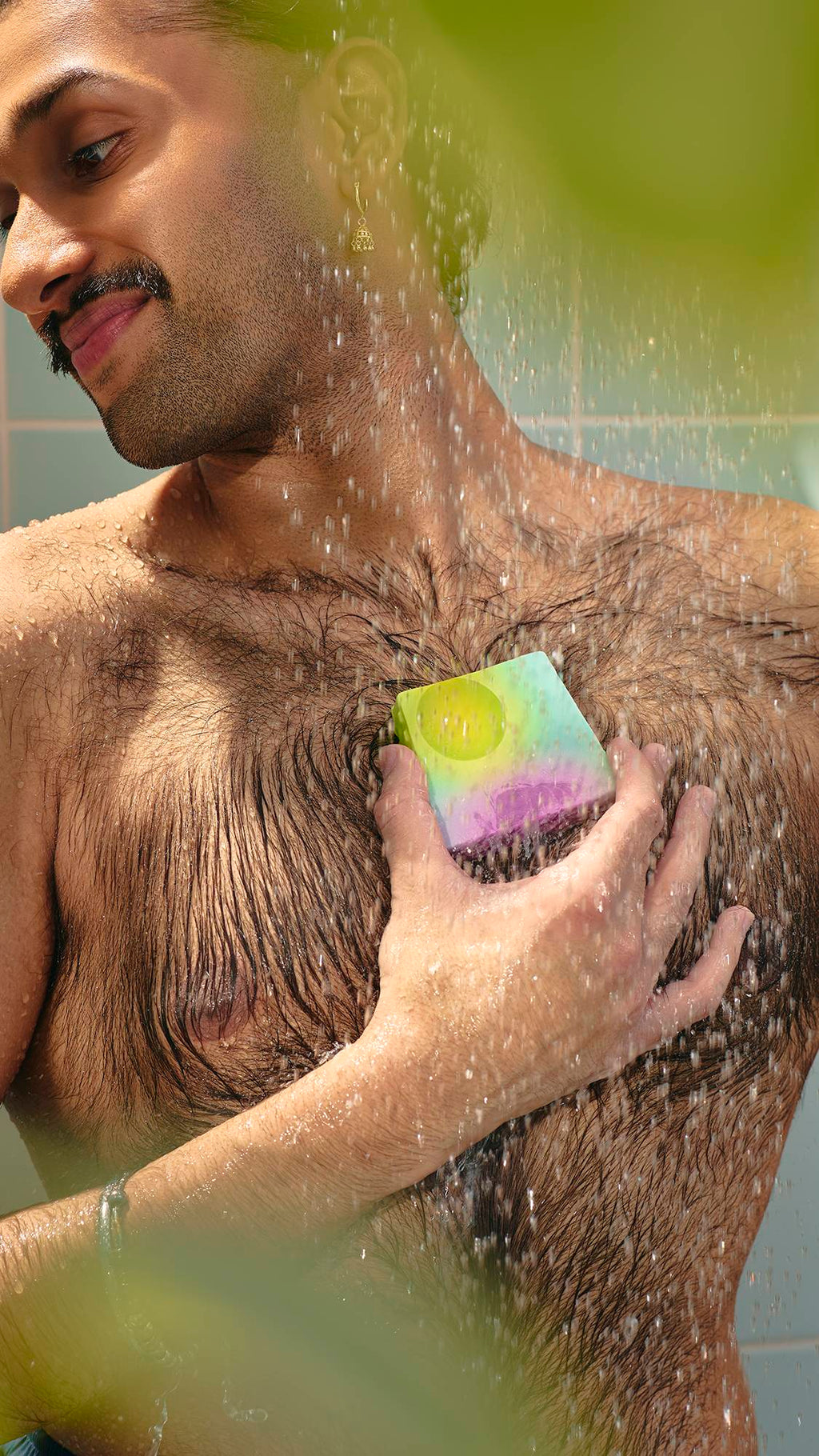 The image shows a model with dark hair in a bun looking down as they lather their chest with a slice of marbled purple and green "To The Moon and Back" soap in the shower. The water creates a fine mist around