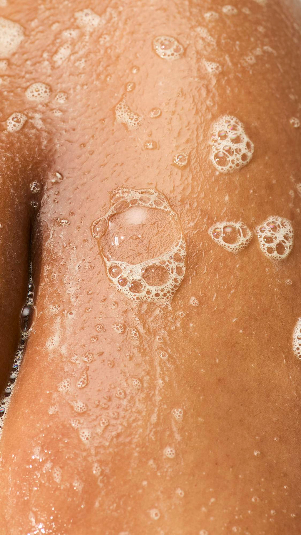 A close-up of wet, glistening skin in the shower, covered in soapy lather and clusters of translucent bubbles.