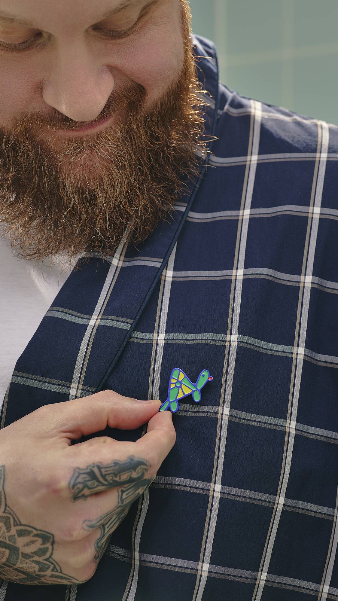 A model with a thick beard and hand tattoos smiles as they pin a small, colourful turtle enamel badge, from the "Turtley Awesome" greeting card, onto the lapel of his navy blue and white checked dressing gown.