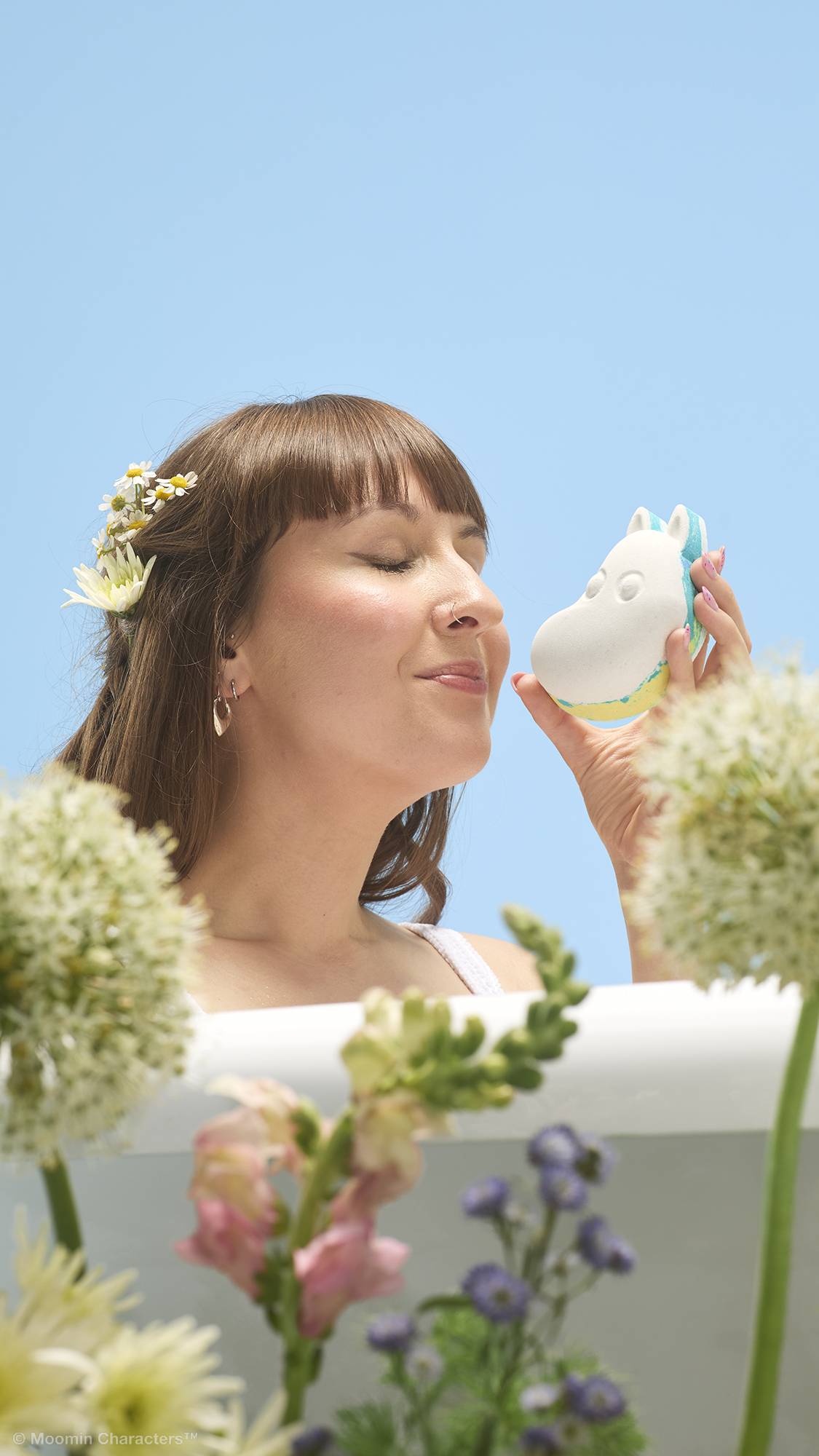 A close-up shot of a person with flowers in their hair, smiling with closed eyes as they bring a white Moomintroll bath bomb up to their nose to smell it. Wildflowers are in the blurry foreground against a clear blue sky.