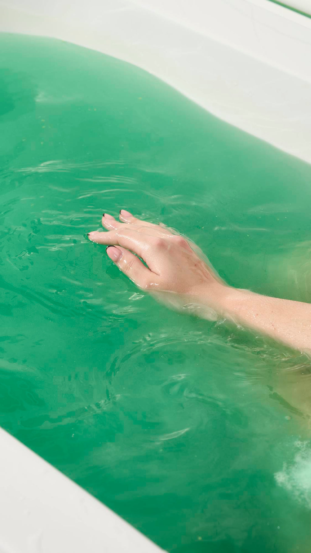 A close-up view of a hand with a pink and black tip manicure resting gently on the surface of vibrant, turquoise-green bathwater. The white tub's edge is visible.