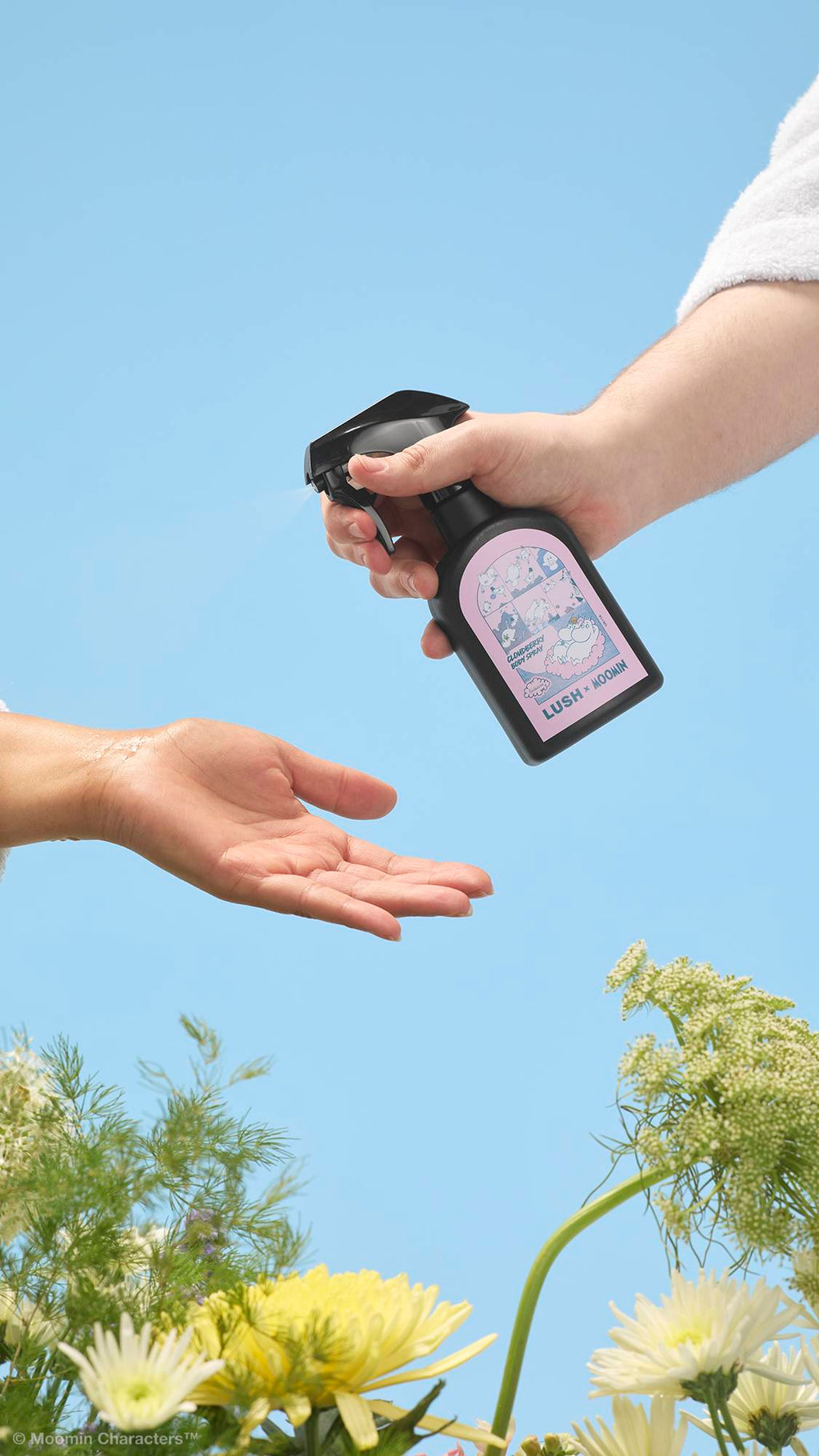 A close-up shot of a hand spraying a black "LUSH x MOOMIN" Cloudberry body spray bottle onto another person's open palm. Yellow and white wildflowers frame the bottom against a clear blue sky background.