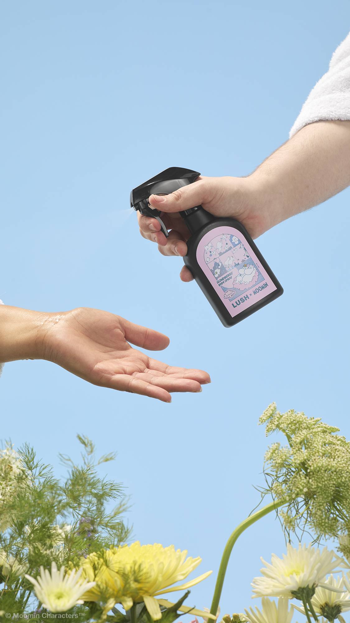 A close-up shot of a hand spraying a black "LUSH x MOOMIN" Cloudberry body spray bottle onto another person's open palm. Yellow and white wildflowers frame the bottom against a clear blue sky background.