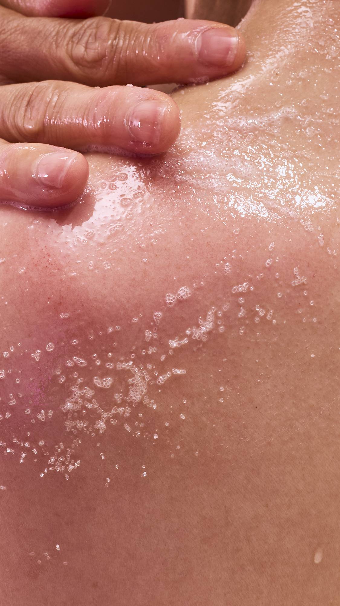 A close-up shot of wet skin with droplets of water and a trail of white, salt-like exfoliating granules. A hand with a pinkish tint is seen at the top, pressing down on the skin as if applying a scrub.