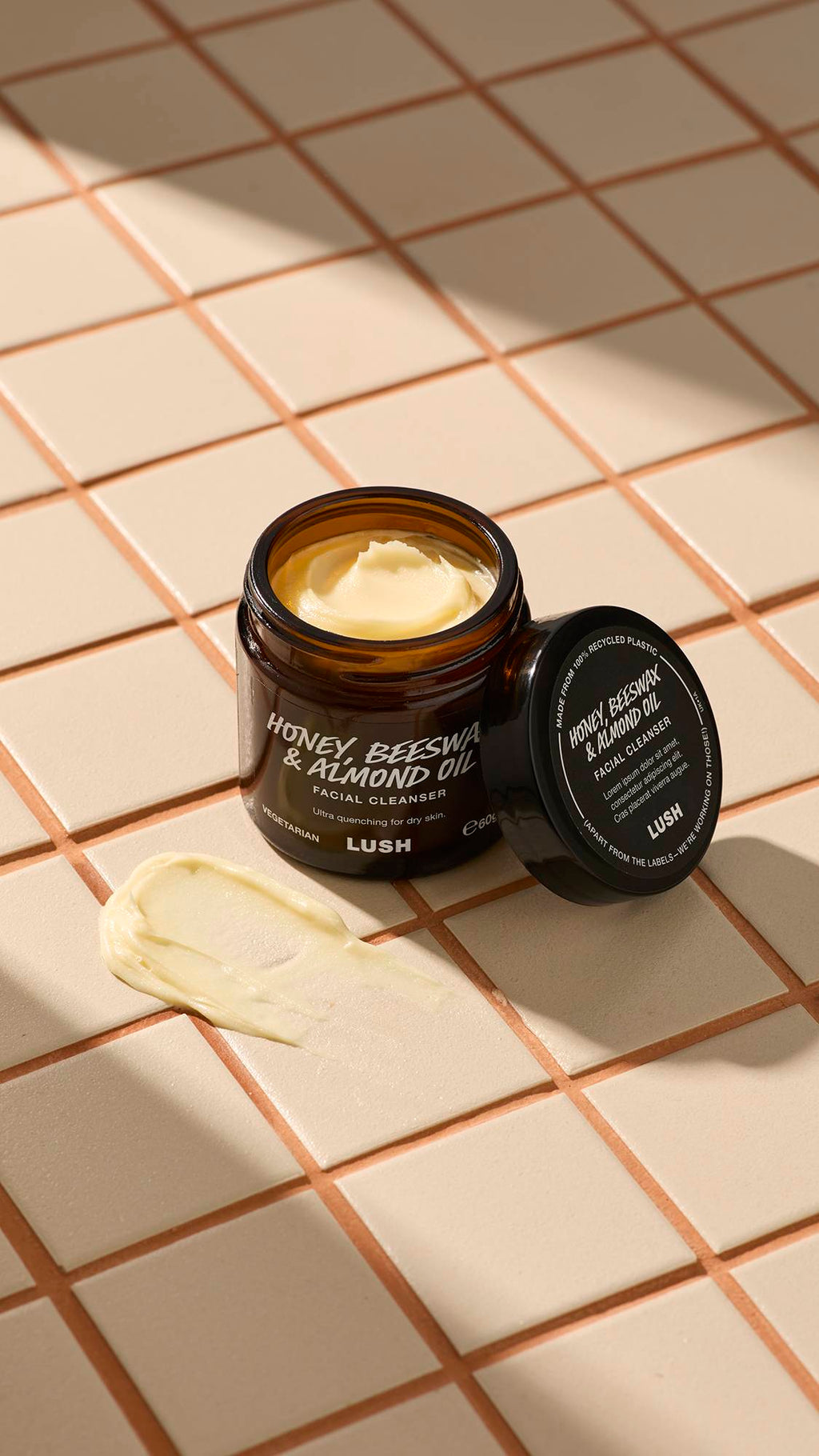 An open brown glass jar of Lush "Honey, Beeswax And Almond Oil" facial cleanser sits on a beige tiled surface. A thick dab of the pale yellow, creamy product is smeared on the tiles next to the jar and its black lid.