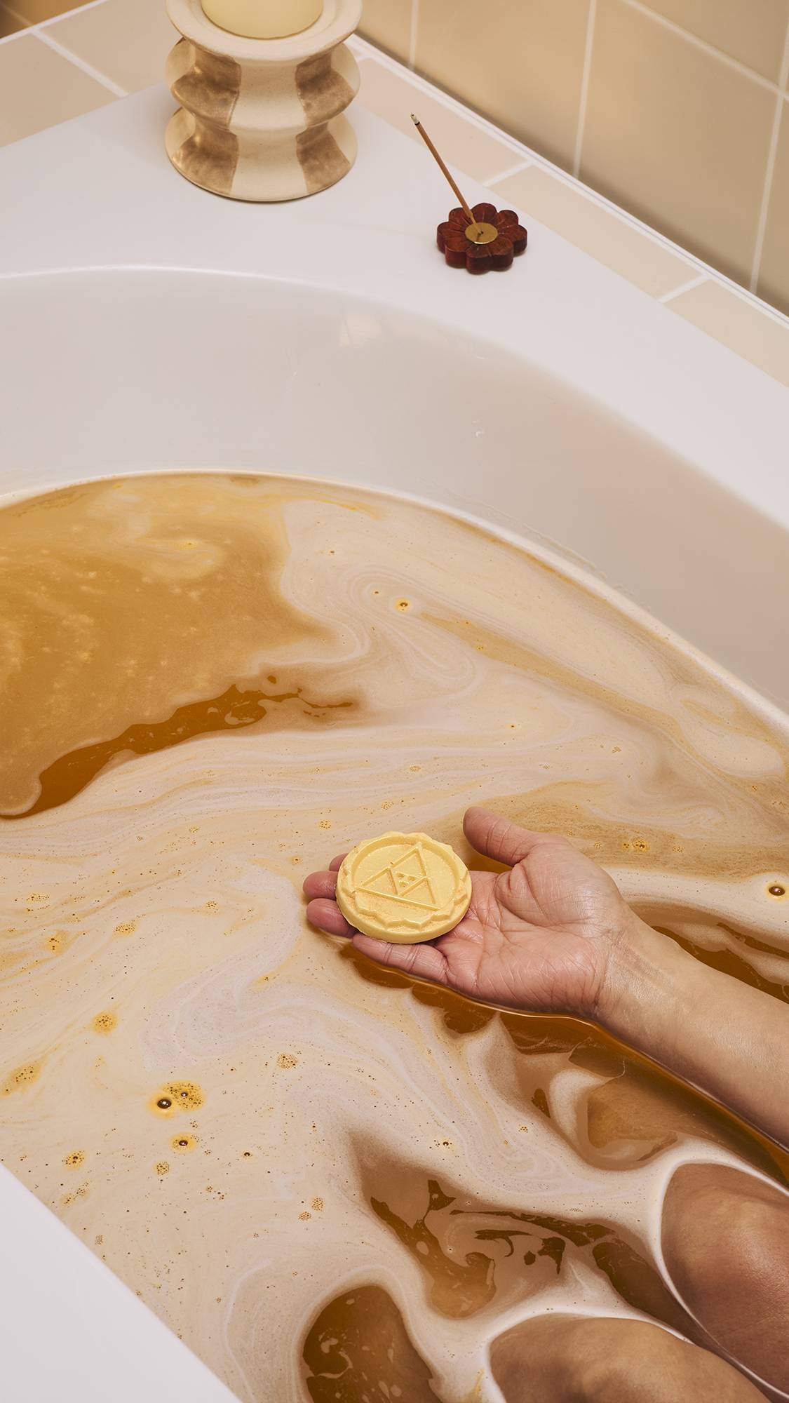 A person rests in a bath filled with amber-coloured, foamy water. They hold a circular yellow solid body oil embossed with a triangle design in their palm. A lit candle and incense stick sit on the bath's edge against neutral-toned tiles.