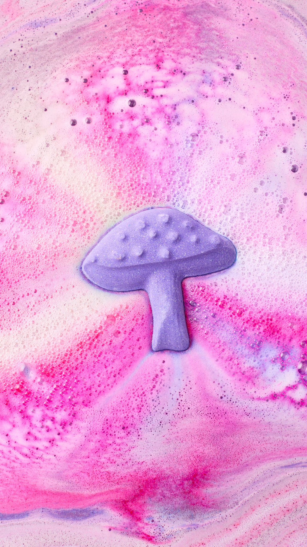An aerial view shows the "Magik Mushroom bath bomb nearly fully dissolved in water, creating a swirling galaxy of pink, violet, and blue foam. The patterns spiral outwards, leaving a trail of thick, colourful bubbles across the water's surface.