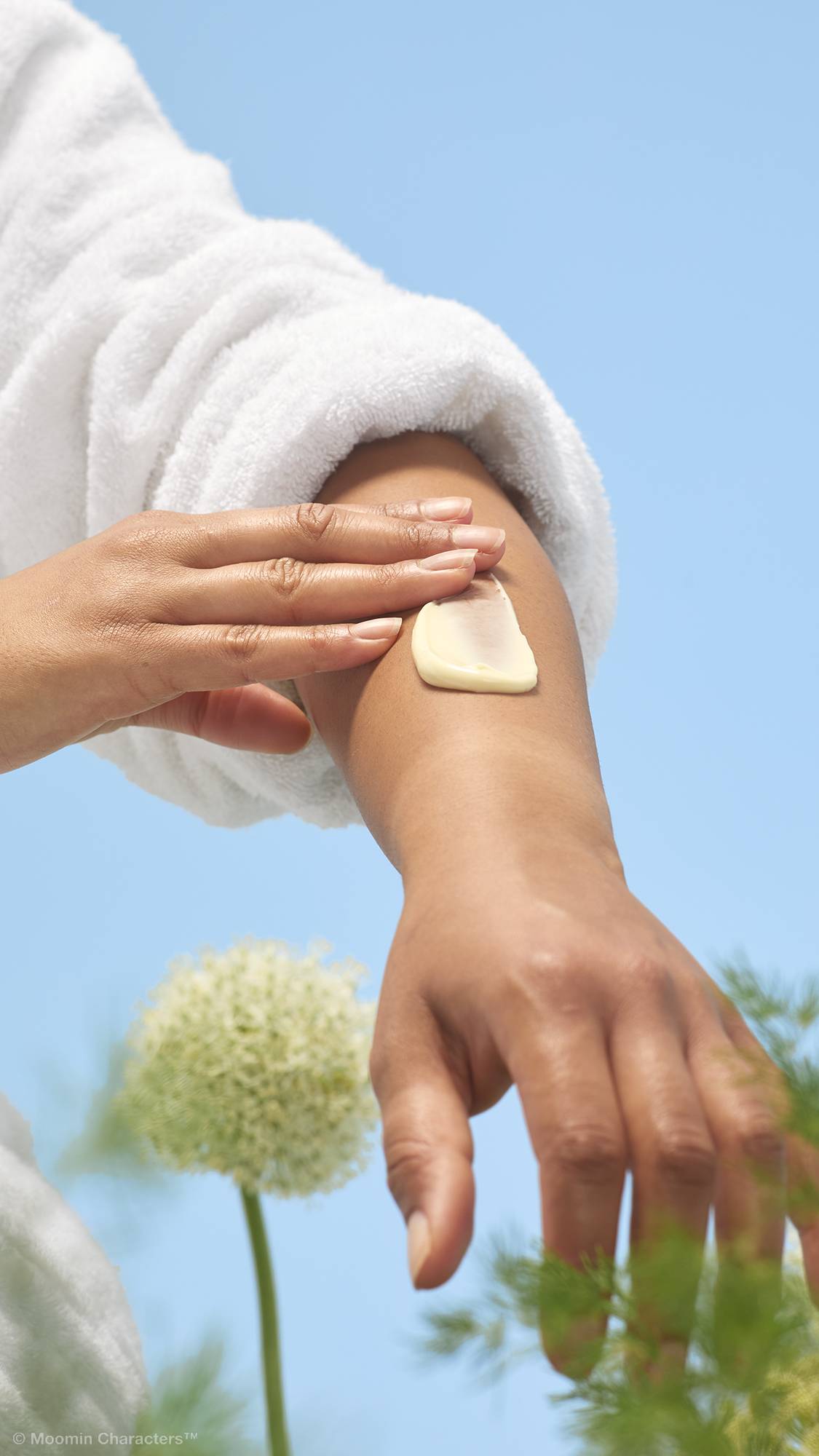 A close-up shot of a person in a white dressing gown smoothing a thick dollop of cream-coloured "Moominmamma" lotion onto their forearm. Soft green foliage and a white flower are visible in the background against a clear blue sky.