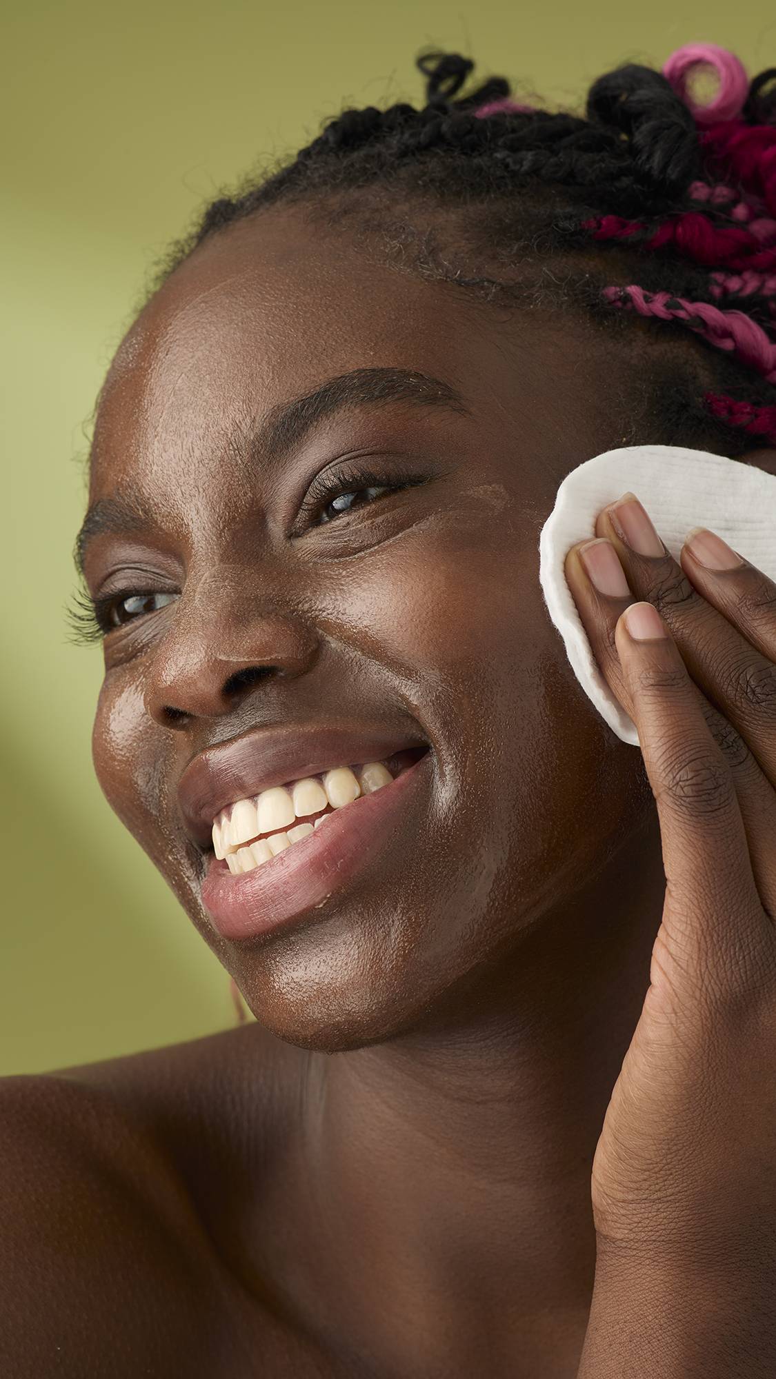 A woman with pink and black braids smiles as she removes the Oat facial cleanser from her face. The thick, creamy product stands out against her skin and the solid olive-green background, emphasising its rich texture.