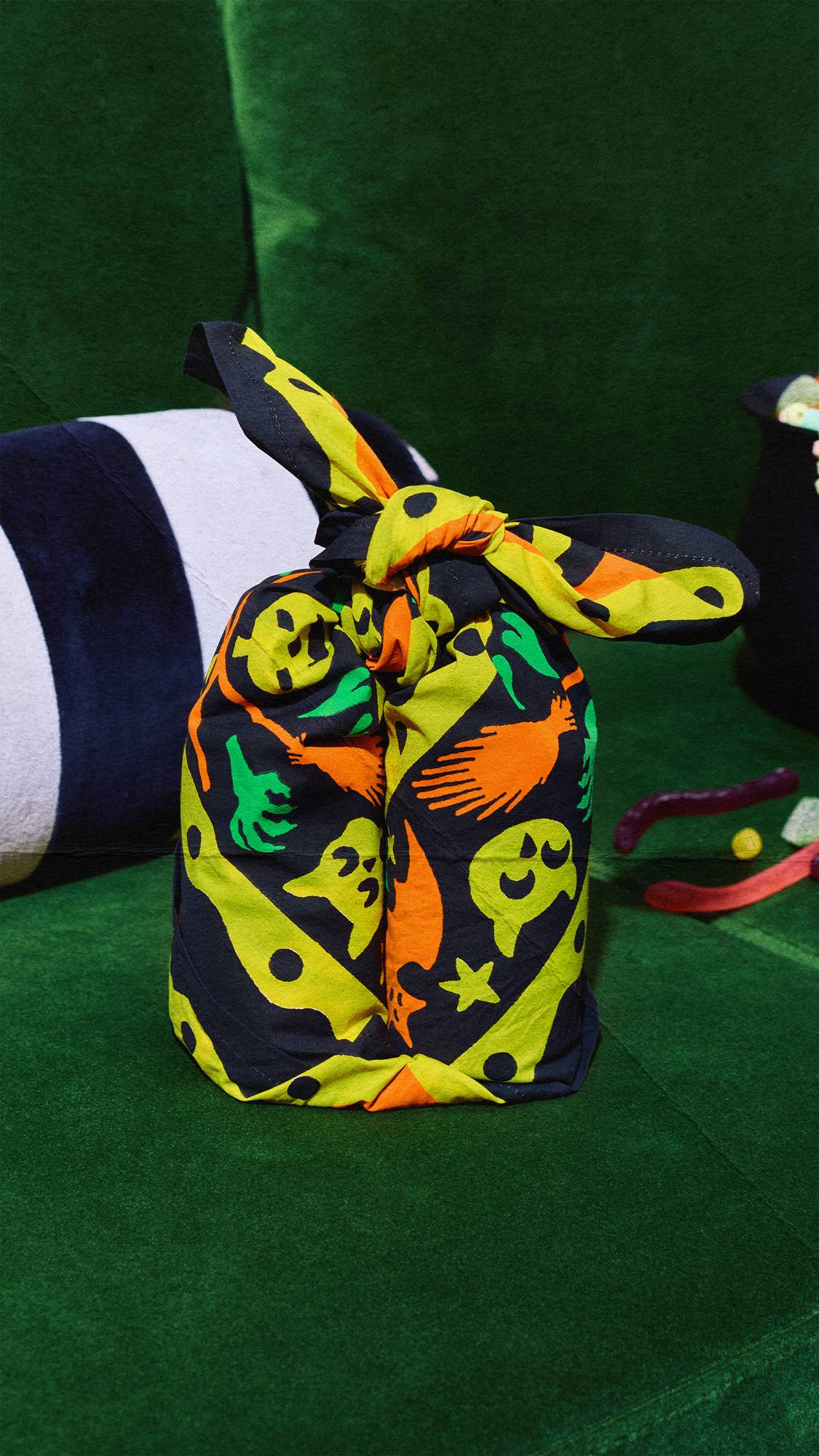 A gift wrapped in a black knot-wrap fabric with a bright lime green and orange Halloween pattern sits on a green velvet couch. The pattern features ghosts, bats, and skulls. Colourful jelly sweets are scattered nearby.