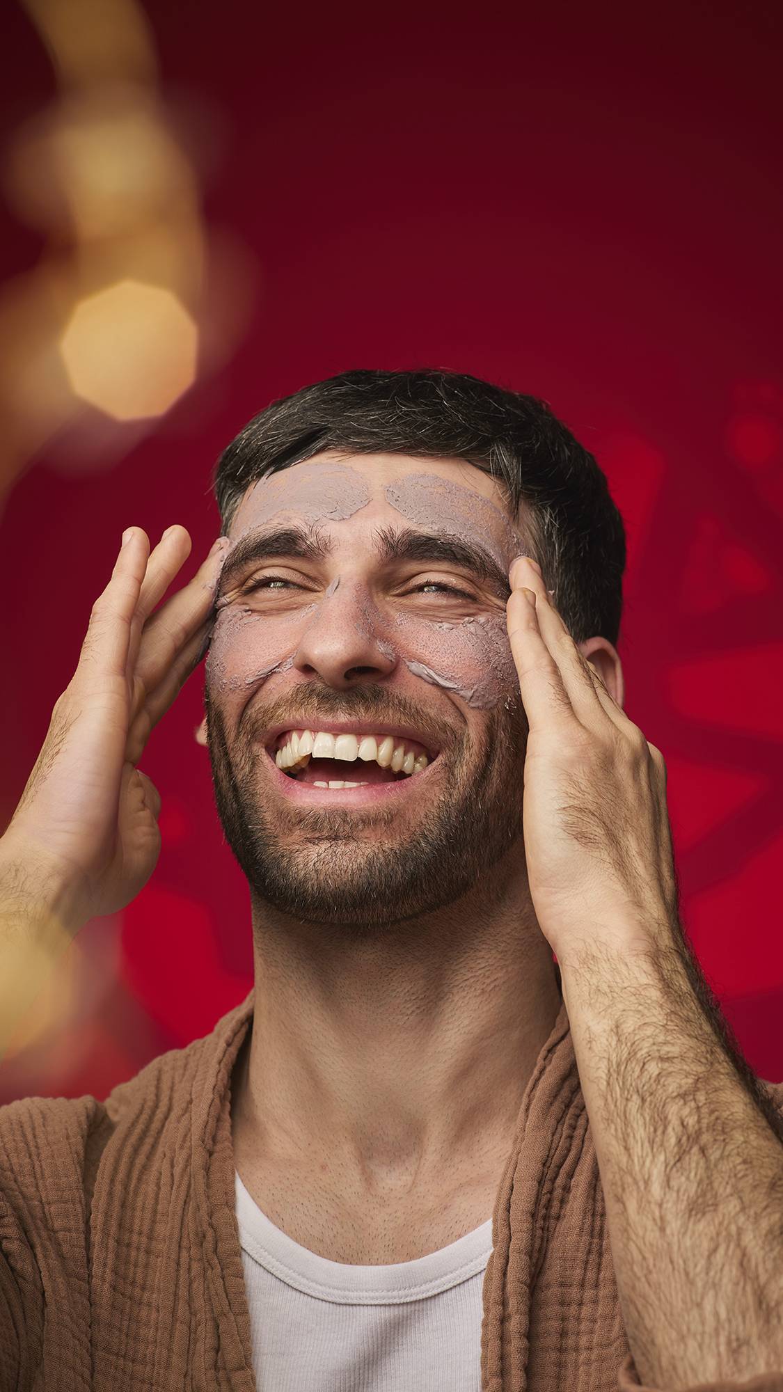 A person laughs while wearing the mauve-brown Sipak face mask across their forehead and cheeks. They wear a brown textured robe against a deep red background with soft golden bokeh lights.