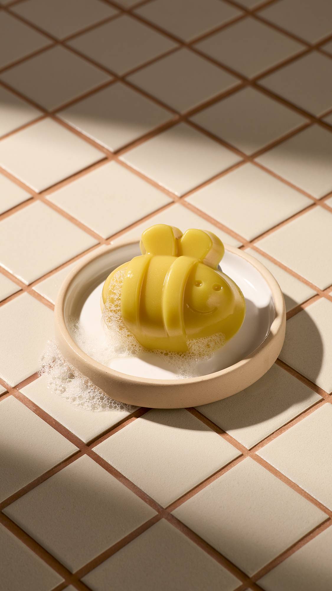 A yellow bee-shaped facial gummy wash with a smiling face rests in a cream-coloured dish. The soap is covered in light white bubbles, and the dish sits on a background of beige square tiles under soft sunlight and shadows.