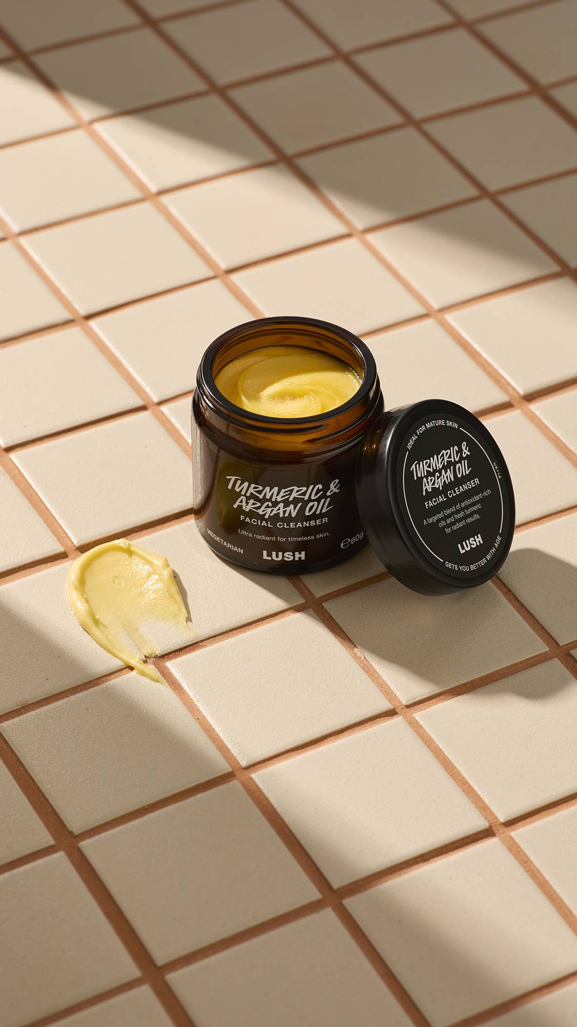 An open brown glass jar of Lush "Turmeric And Argan" facial cleanser sits on a beige tiled surface. A thick dab of the pale yellow, creamy product is smeared on the tiles next to the jar and its black lid.