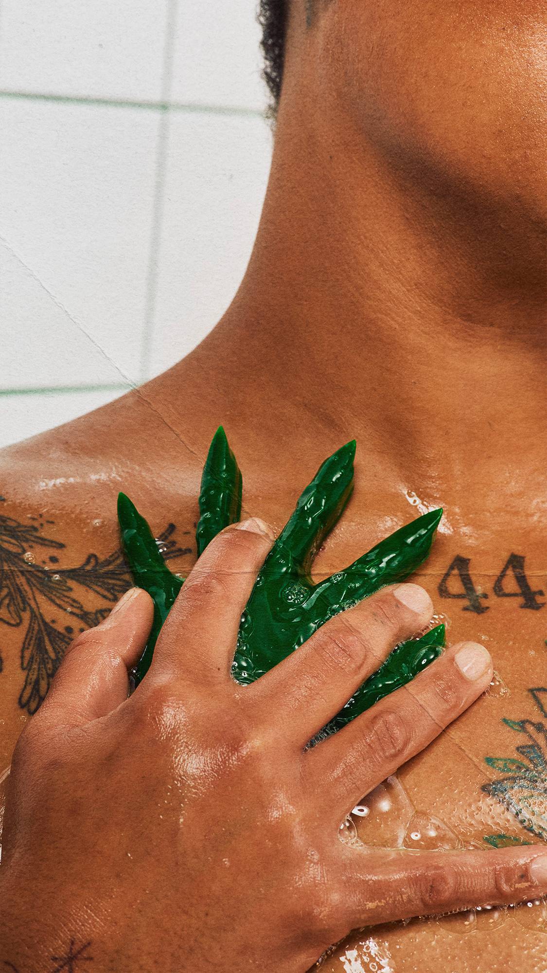 A close-up of a person using a dark green shower gummy shaped like a witch's hand to wash their tattooed chest in the shower. One hand presses the soapy, textured bar against wet skin. White square wall tiles with green grout form the background.