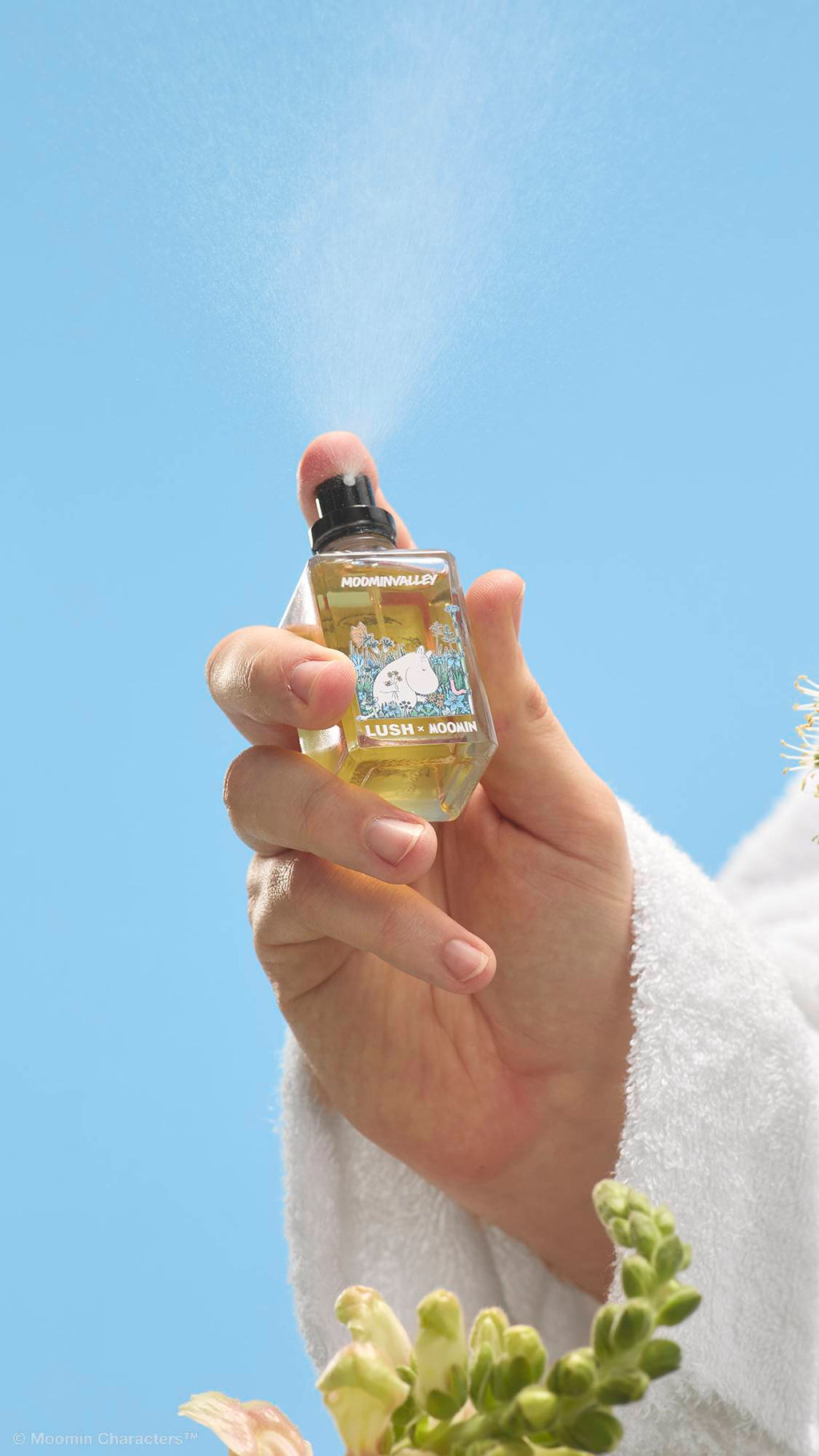 A close-up shot of a hand in a white dressing gown pressing the nozzle of a Lush x Moomin perfume bottle, releasing a fine mist into the air. Wildflowers are visible in the foreground against a clear blue sky.