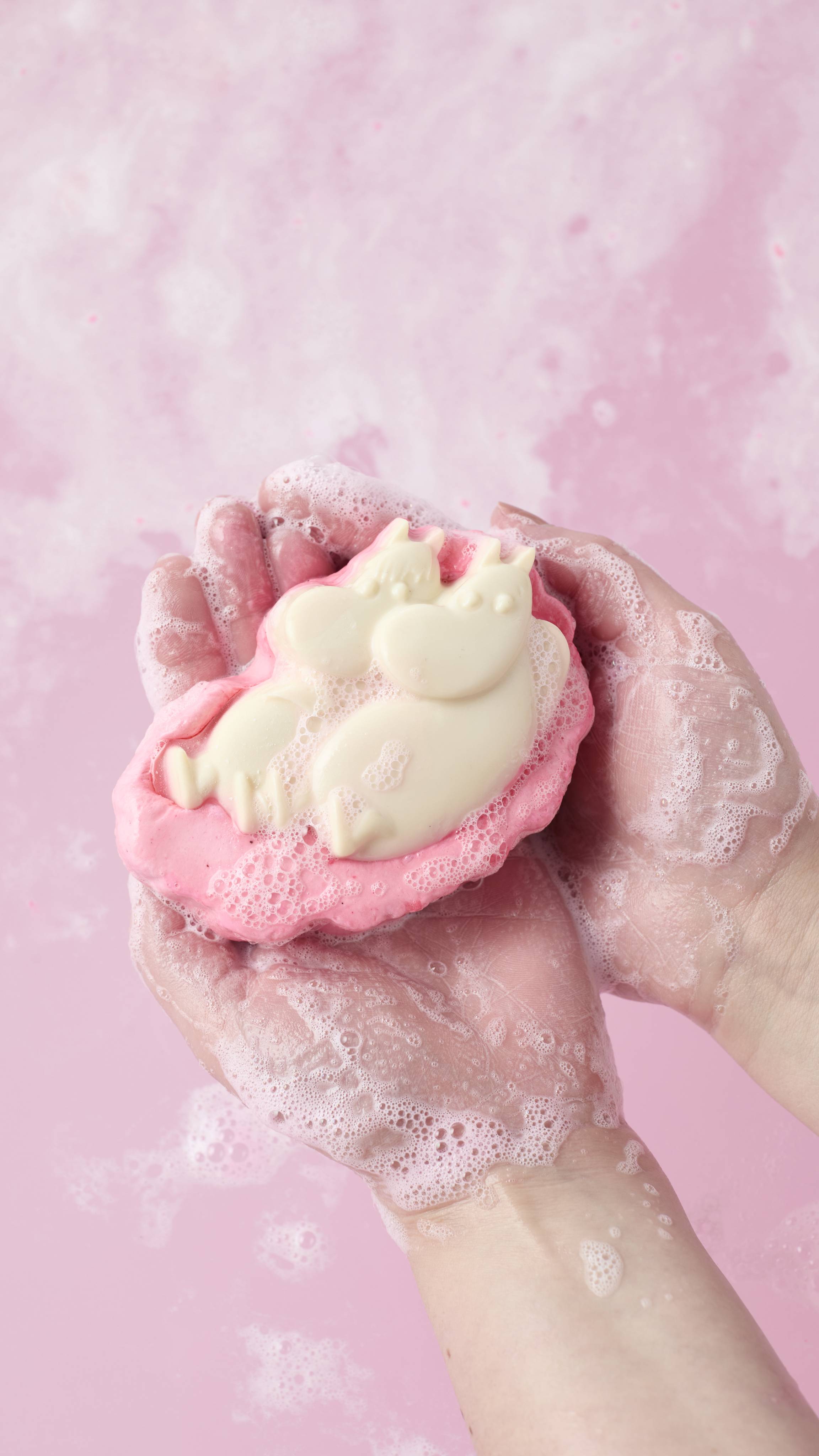 An overhead shot of two soapy hands cradling a pink and cream-coloured "Moomin Delight" soap and body wash product. White foam covers the skin, set against a background of swirling, bubbly pink water.