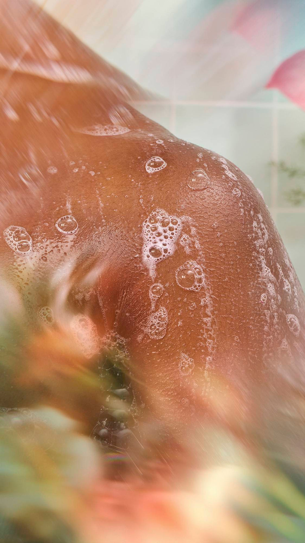 A close-up shows water streaming down a wet shoulder, with small soapy bubbles clinging to the skin. The scene is framed by blurred pink and green floral elements against a mint green tiled wall.
