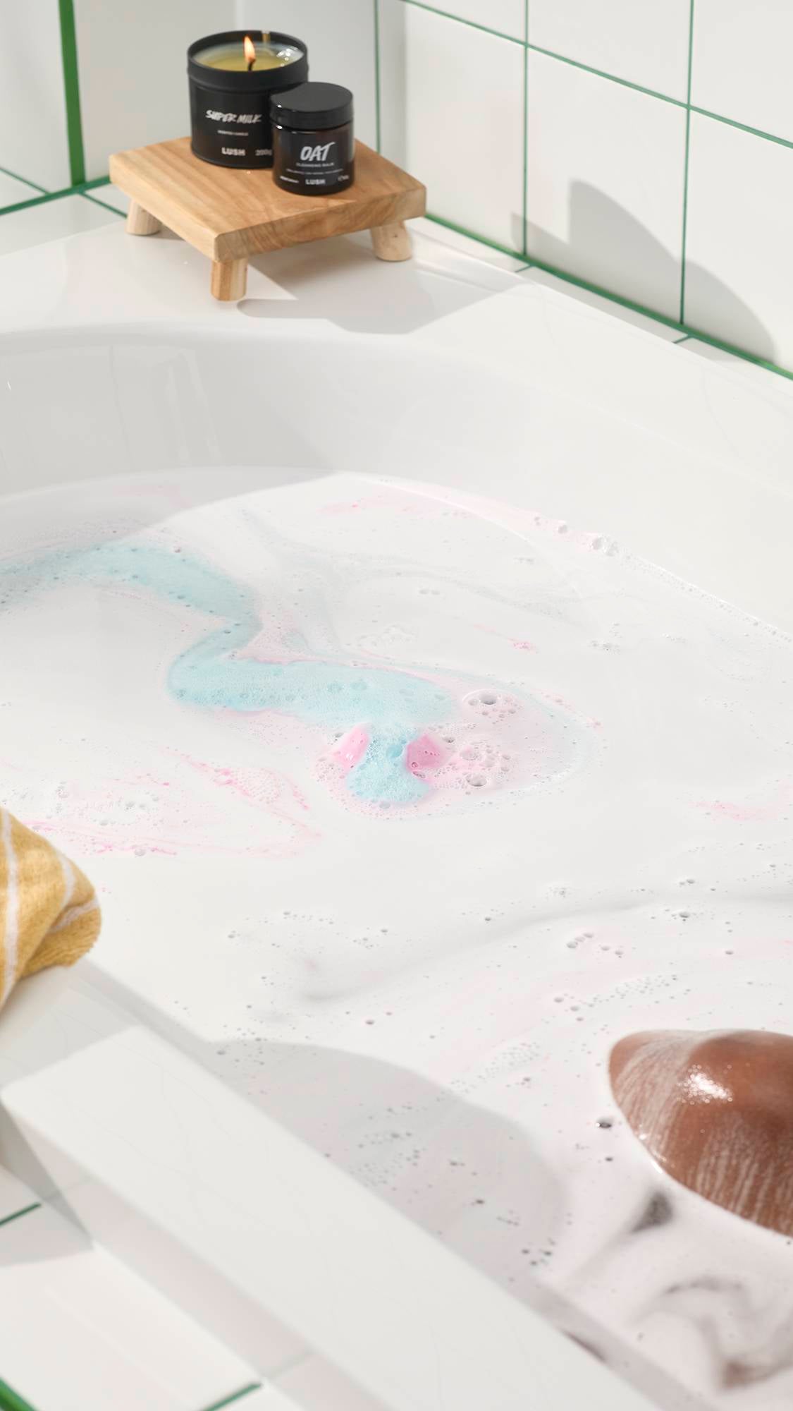 Bathtub filled with milky white water and swirls of pink and blue foam from a Twilight Bath Bomb. A person relaxes in the tub while a candle and jar sit on a wooden stand nearby.