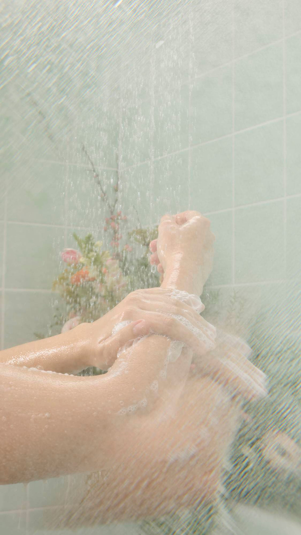 Wet, soapy hands massage "Crème Brûlée" Body Scrub onto an arm under a shower stream. The lathering action takes place against a background of mint green tiles and soft-focus flowers.