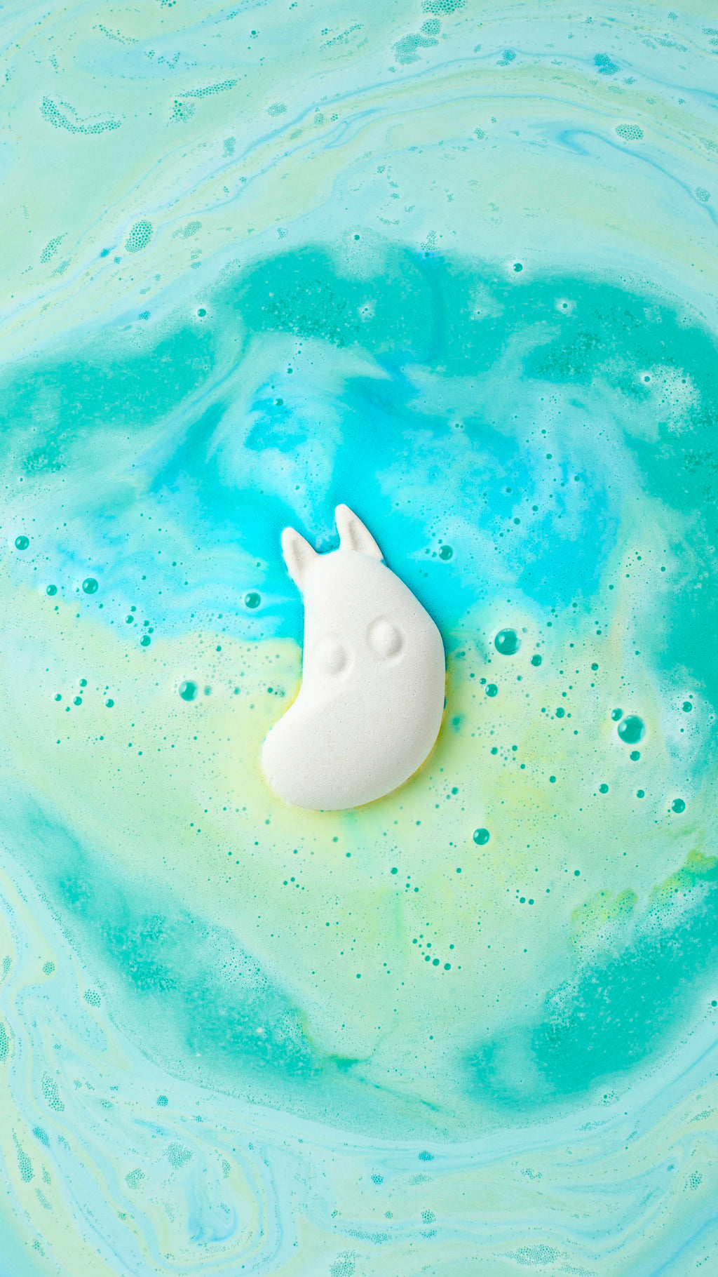 An overhead shot of a white Moomintroll bath bomb dissolving in water, viewed from a slightly different angle. It floats amidst swirling trails of turquoise, blue, and yellow foam, creating a marbled pattern across the bubbling surface.