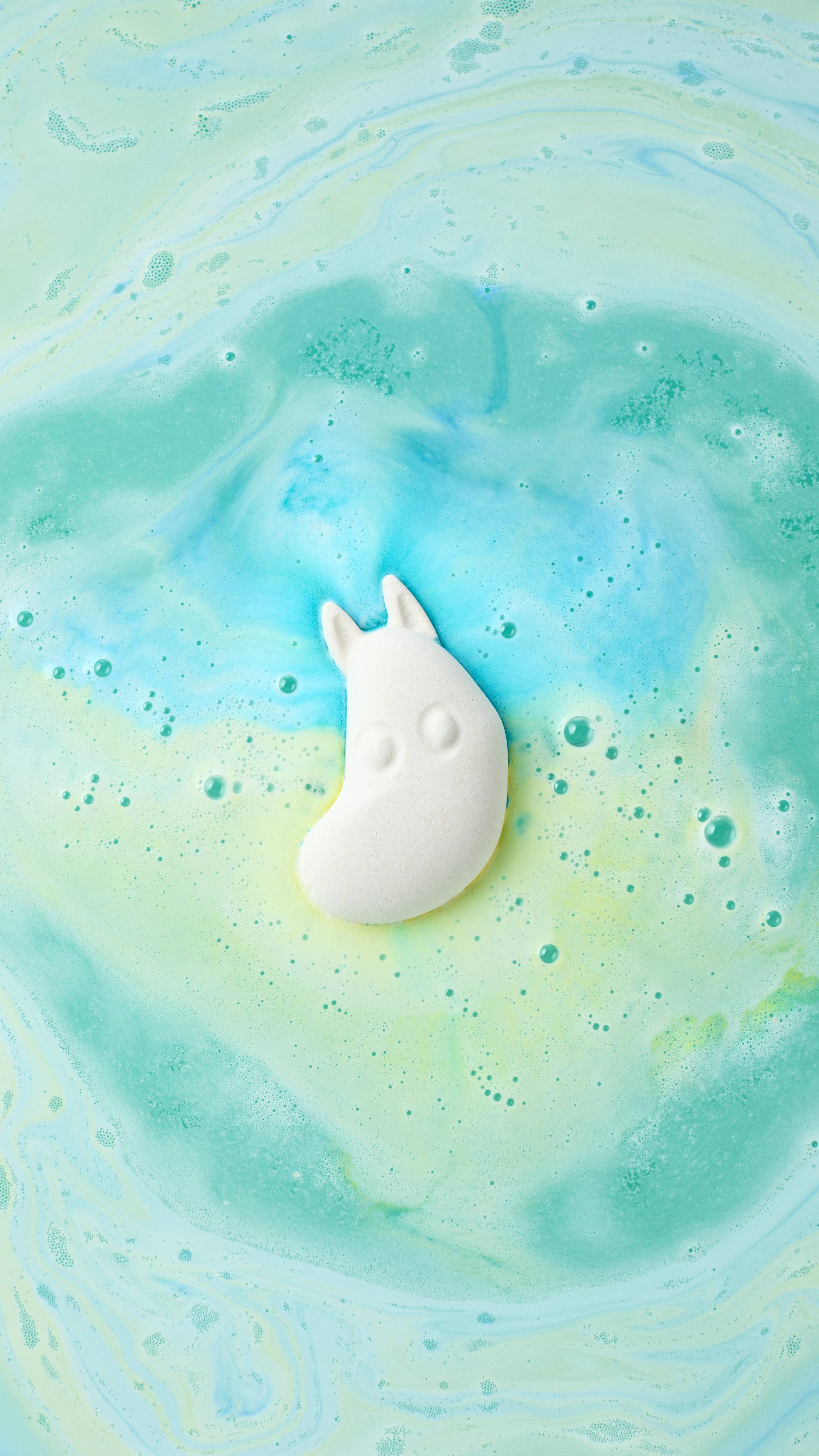 An overhead shot of a white Moomintroll bath bomb dissolving in water, viewed from a slightly different angle. It floats amidst swirling trails of turquoise, blue, and yellow foam, creating a marbled pattern across the bubbling surface.