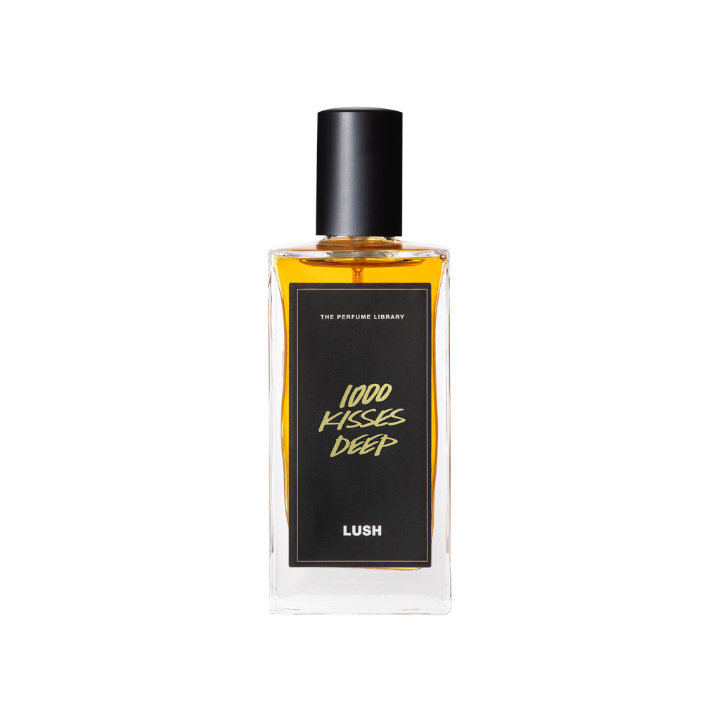 A glass perfume bottle filled with amber liquid. A black label reads 'The Perfume Library' and '1000 Kisses Deep'.
