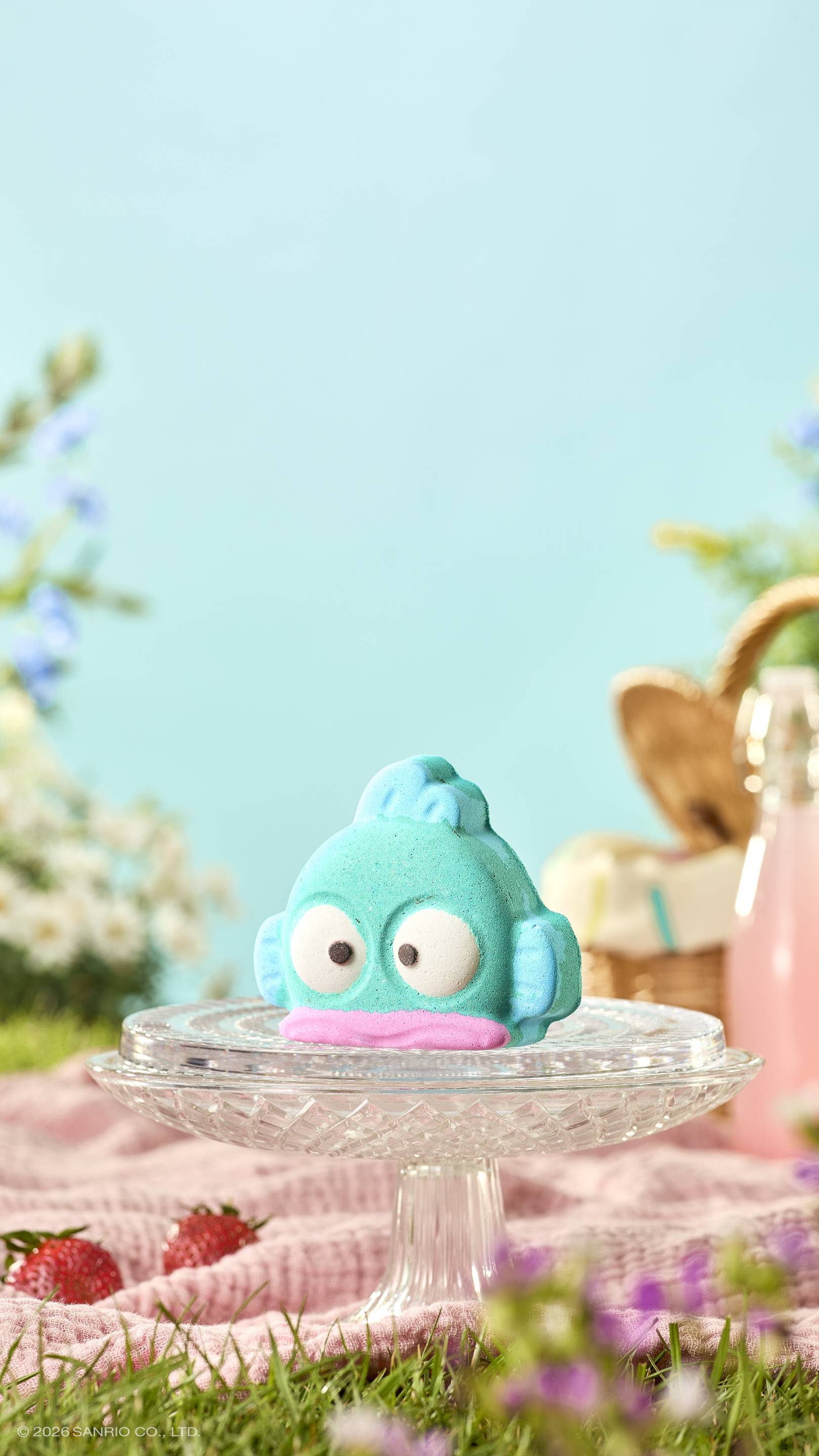 The teal, fish-shaped "Hangyodon" bath bomb sits on a glass cake stand in a sunny picnic setting. The background is a blue sky, with a wicker basket and a drink out of focus. A pink blanket and strawberries are scattered on the grass below.