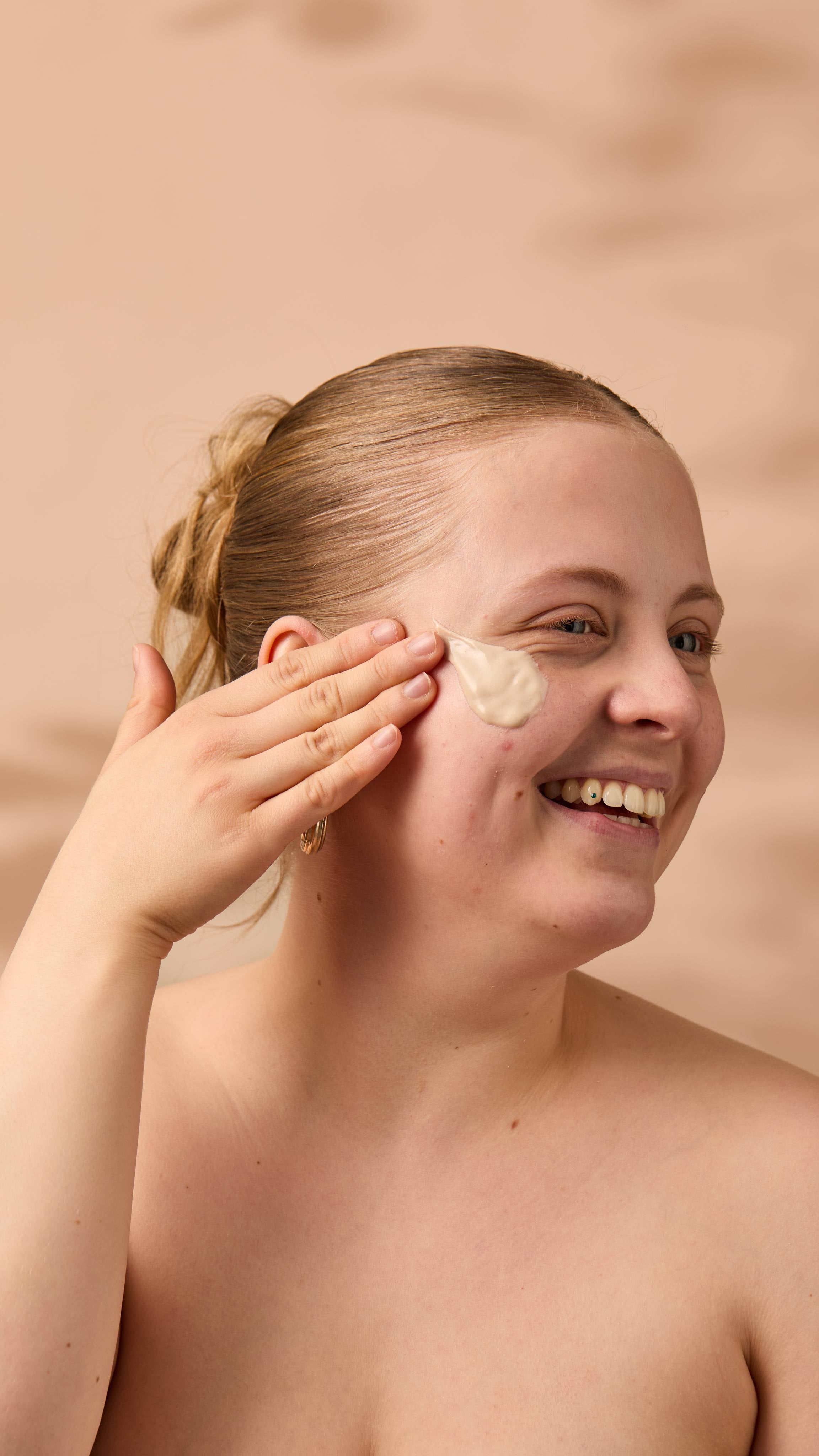 An image of the model's face on a warm, beige background. Their copper hair is tied back, and they have applied the thick Coconut Shie face mask onto their cheek as they smile.