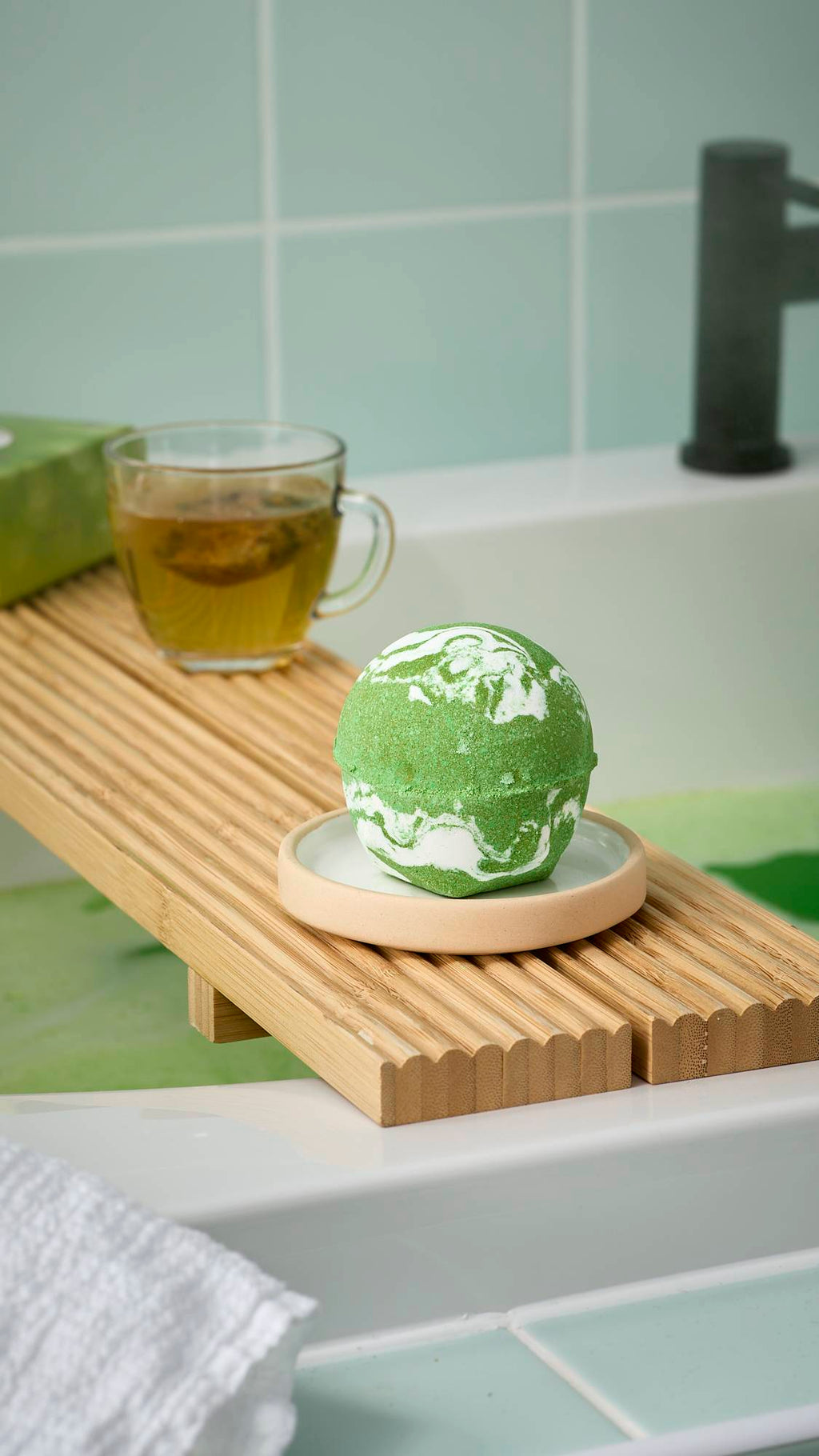 The green and white marbled "Cold Bath Bomb" rests on a wooden bath tray alongside a cup of tea. Green bath water is visible below, framed by mint green tiles and a white towel in the foreground.