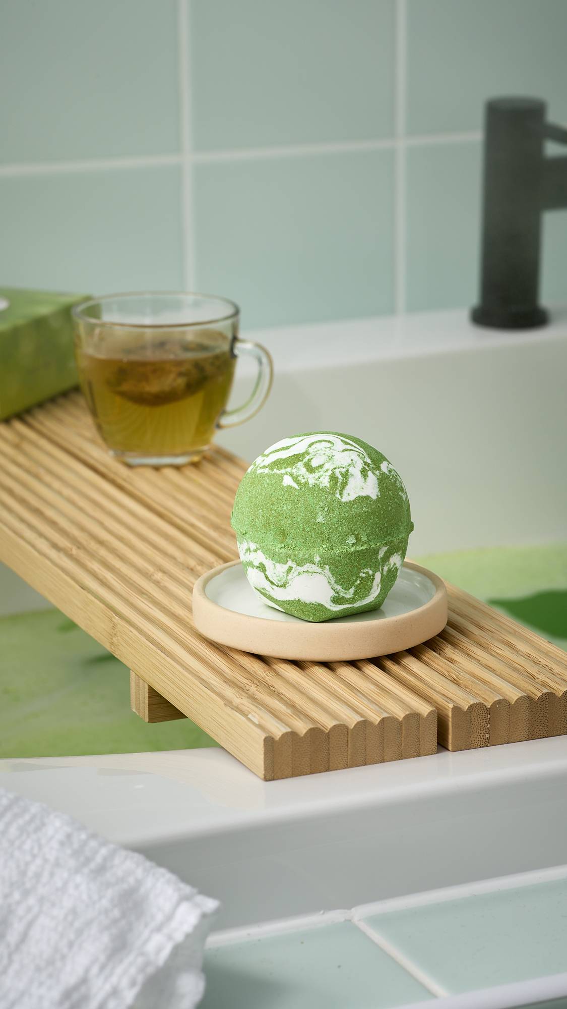 The green and white marbled "Cold Bath Bomb" rests on a wooden bath tray alongside a cup of tea. Green bath water is visible below, framed by mint green tiles and a white towel in the foreground.