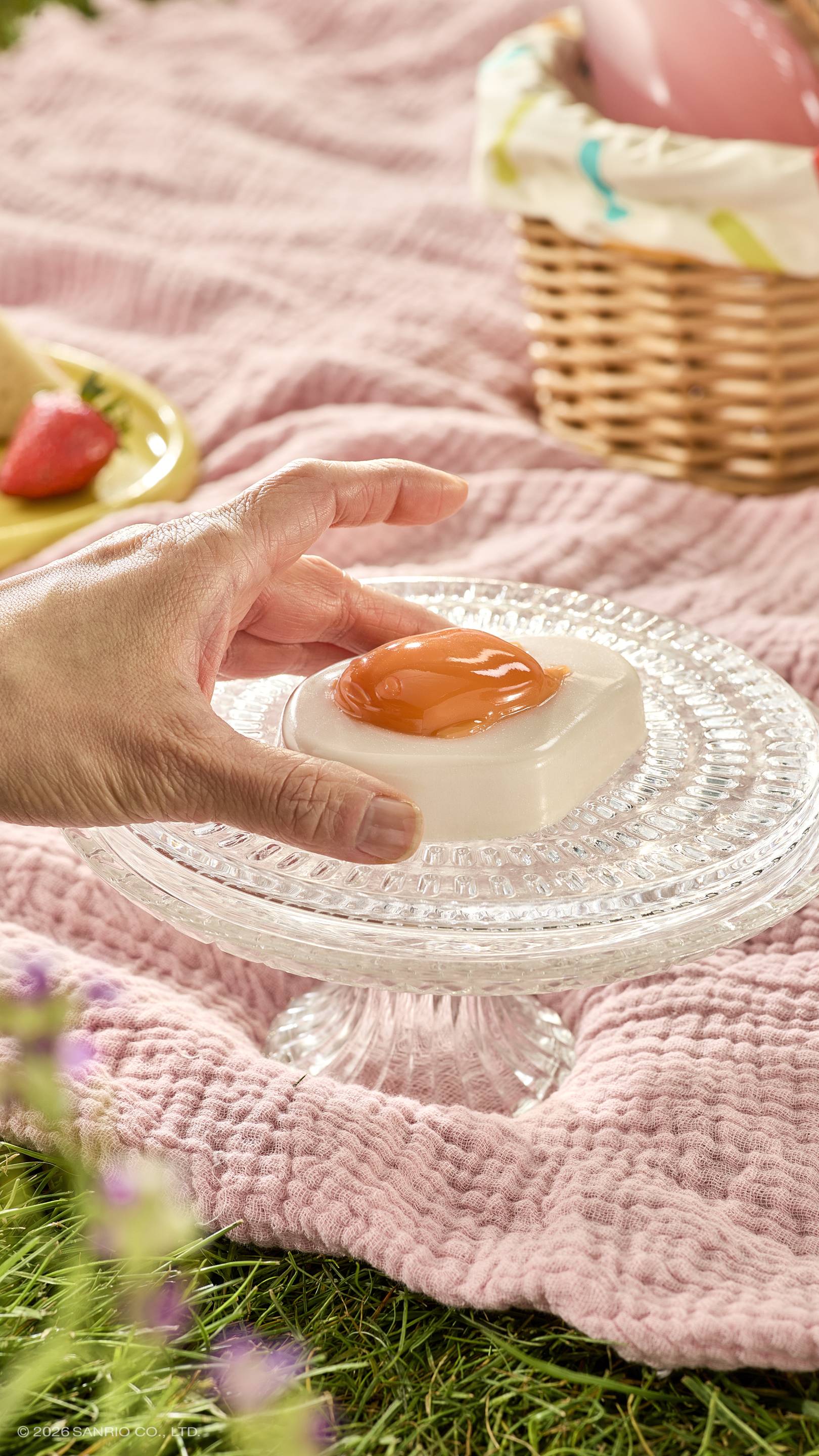 A hand reaches for a glossy fried-egg-shaped shower jelly sitting on a glass cake stand. The sunny picnic scene features a pink blanket on the grass, a wicker basket, and a strawberry.