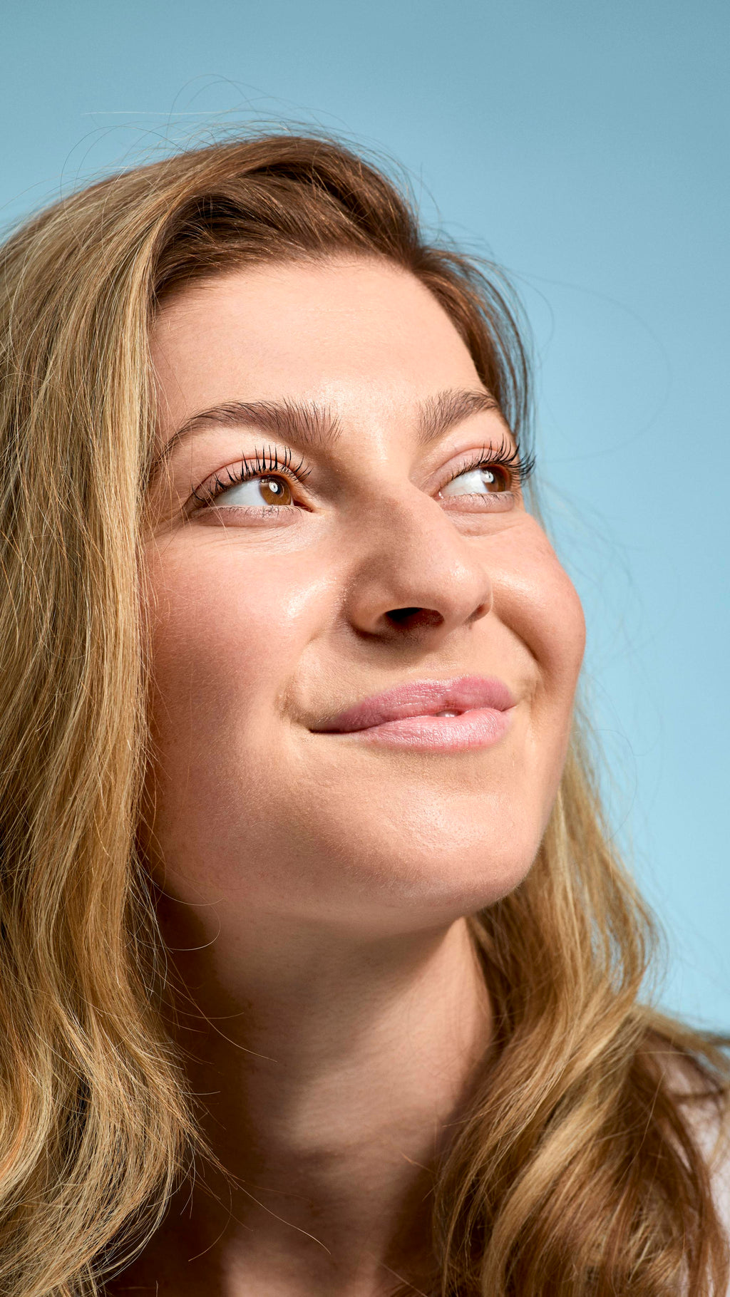 A close-up of a model with fair skin and dark blonde hair looking upwards with a soft, natural smile, gently pouting freshly balmed lips. Their face has a radiant, dewy finish, set against a simple, light-blue background.