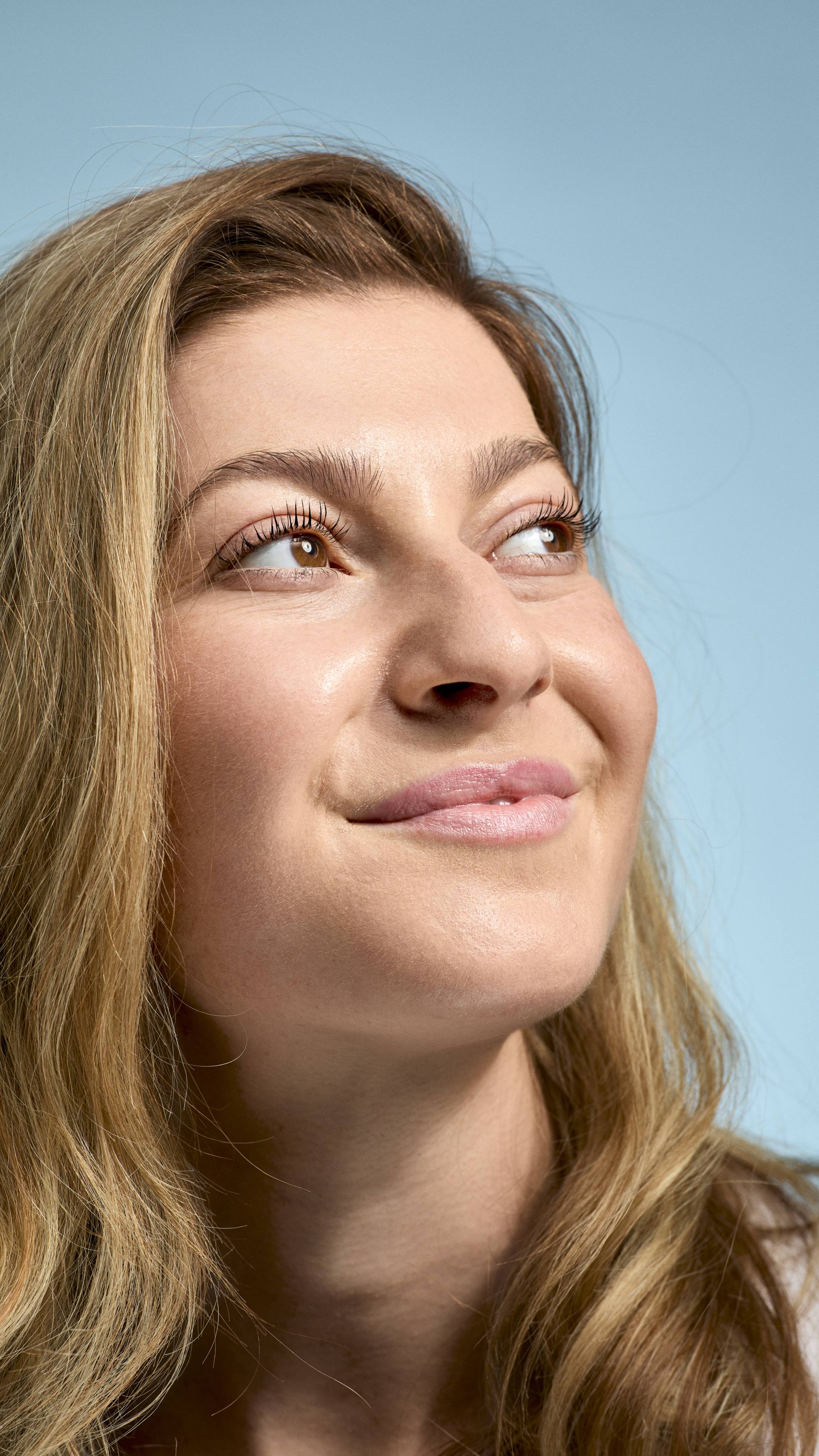A close-up of a model with fair skin and dark blonde hair looking upwards with a soft, natural smile, gently pouting freshly balmed lips. Their face has a radiant, dewy finish, set against a simple, light-blue background.